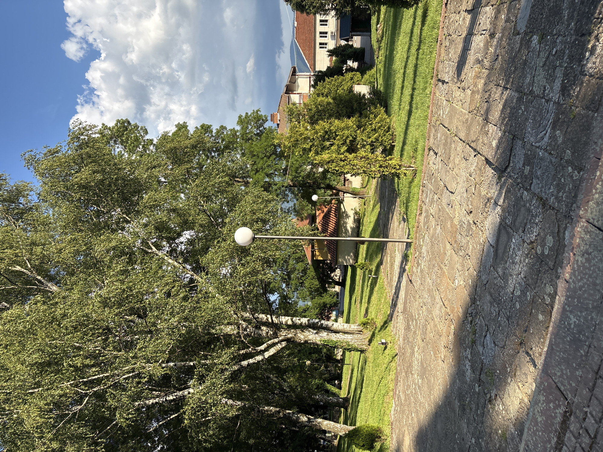 The image shows a park-like setting with trees, grass, and paved walkways. In the foreground, there is a stone pathway with shadows cast by the trees. To the right, a lamppost with a spherical light fixture stands on the grass. In the background, there are several buildings with red tile roofs, and lush green trees surround the area. The sky is blue with scattered white clouds, indicating daytime. The location appears to be a public space, possibly a garden or a courtyard associated with one of the buildings. There are no people visible in the image. The overall impression is one of a well-maintained outdoor area with a mix of natural and architectural elements.
