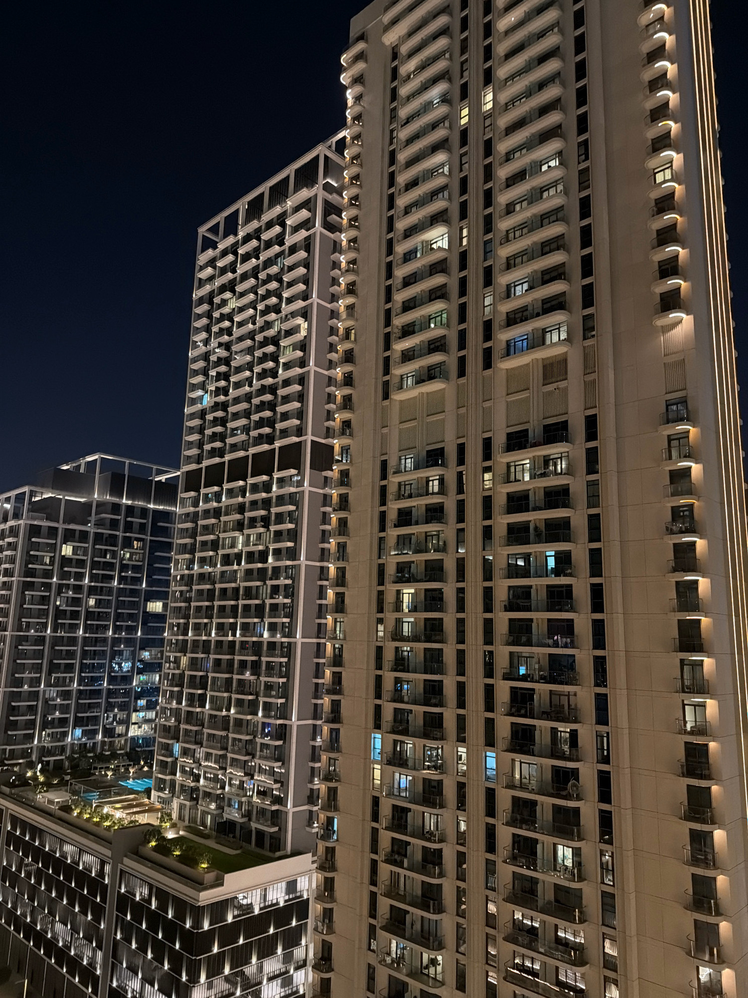 The image shows a nighttime view of the modern architecture of Dubai, United Arab Emirates. The scene is dominated by two tall residential buildings. The building in the foreground is a light-colored, high-rise structure with many balconies and rows of illuminated windows. Vertical light strips accentuate the building's height. To the left, a second, darker-colored building is visible, also featuring numerous balconies and lit windows, with a lower section that includes a rooftop pool and landscaped area. The sky above is dark, indicating nighttime. There are no people or overt actions depicted, but the illuminated windows suggest occupancy. The overall impression is one of urban development and nighttime city life.