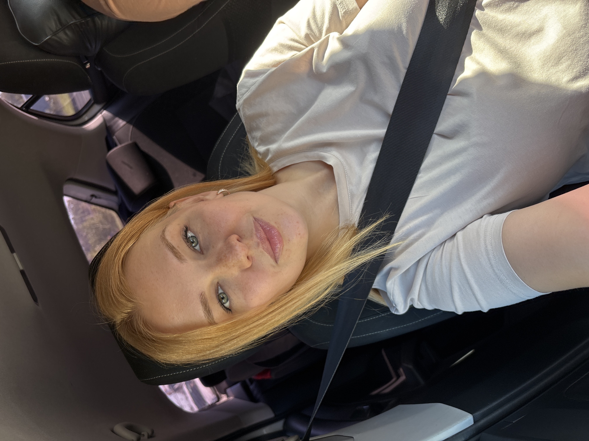 A fair-skinned woman with strawberry blonde hair and green eyes, wearing a light beige t-shirt and a seatbelt, is seen from the chest up. She has a smattering of freckles across her nose and cheeks. The woman is seated in the passenger seat of a car, with the interior visible behind her. The sunlight streams in from the right, illuminating her face and t-shirt. The background includes car seats and a window showing a blurred outdoor scene. The image appears to be a selfie, taken during the daytime. The location is identified as Zhukovo, Russia.