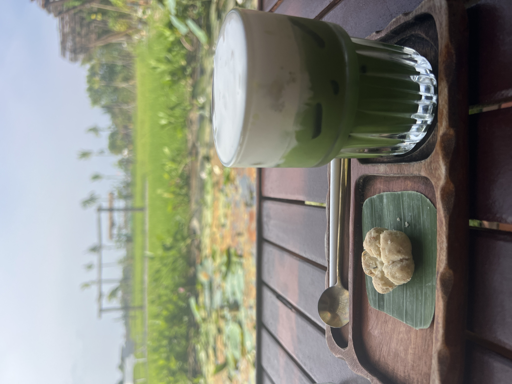 A close-up shot captures a refreshing glass of matcha green tea with a creamy white foam topping, accompanied by a small cookie on a banana leaf and a spoon, all presented on a wooden tray. The tray rests on a rustic wooden table. In the background, a serene natural landscape unfolds, featuring lush green rice fields, a pond with water lilies, and a glimpse of traditional Vietnamese architecture, suggesting the location is a cafe or restaurant in Hoi An, Vietnam, offering a tranquil outdoor dining experience. The sky is overcast, casting a soft light on the scene.