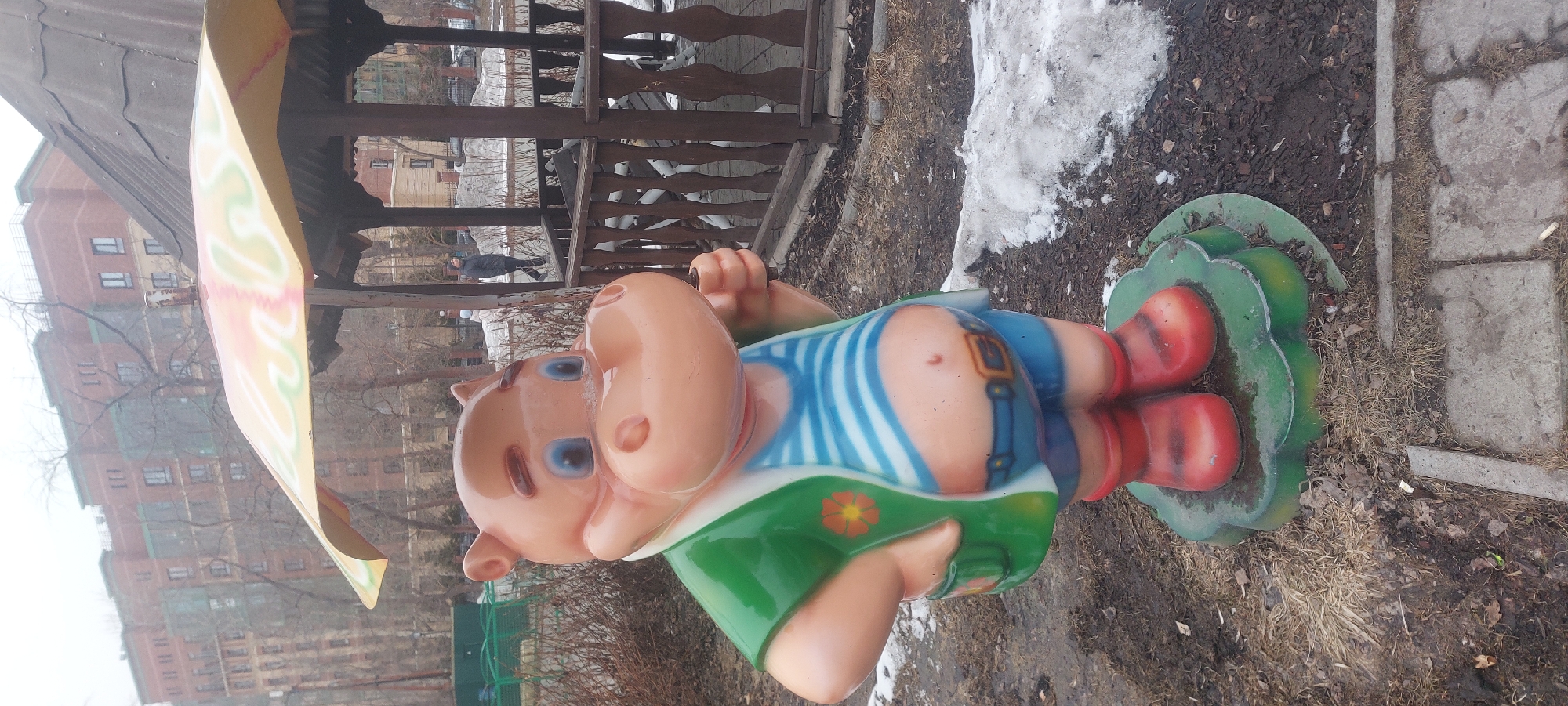 The image shows a brightly colored hippopotamus statue in Novosibirsk, Russia. The statue is of a cartoonish hippopotamus wearing a striped shirt, blue shorts with a belt, red boots, and a green apron with a flower. It is placed on a green, flower-shaped base next to a patch of melting snow and dry ground with some grass and debris. The background includes a wooden gazebo, a multi-story building, bare trees, and a cloudy sky. A person in dark clothing can be seen walking past the gazebo. The statue's colors and style suggest it is a decoration in a park or playground.