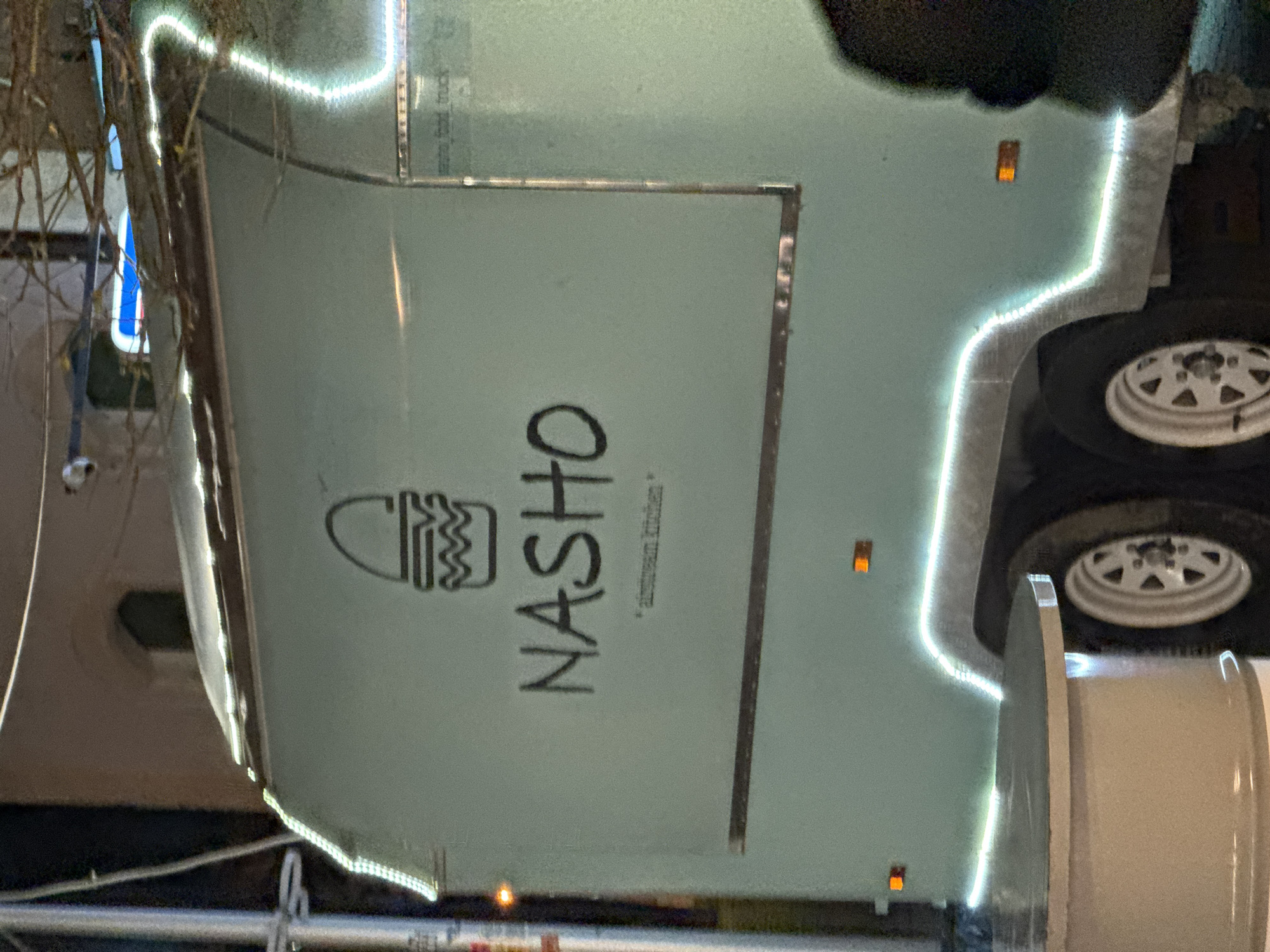 Here is a description of the image:
The image shows a light green food truck with the name "NASHO" in black lettering on the side. A logo resembling a burger is to the left of the name. Below the name it reads "airstream kitchen." The truck is outlined with white LED lights. A wheel and some equipment are visible on the right side of the frame. Some foliage and a building are visible in the background on the left side of the image. The image is rotated 90 degrees counterclockwise. I cannot determine the specific location in Varna, Bulgaria, from the image alone.