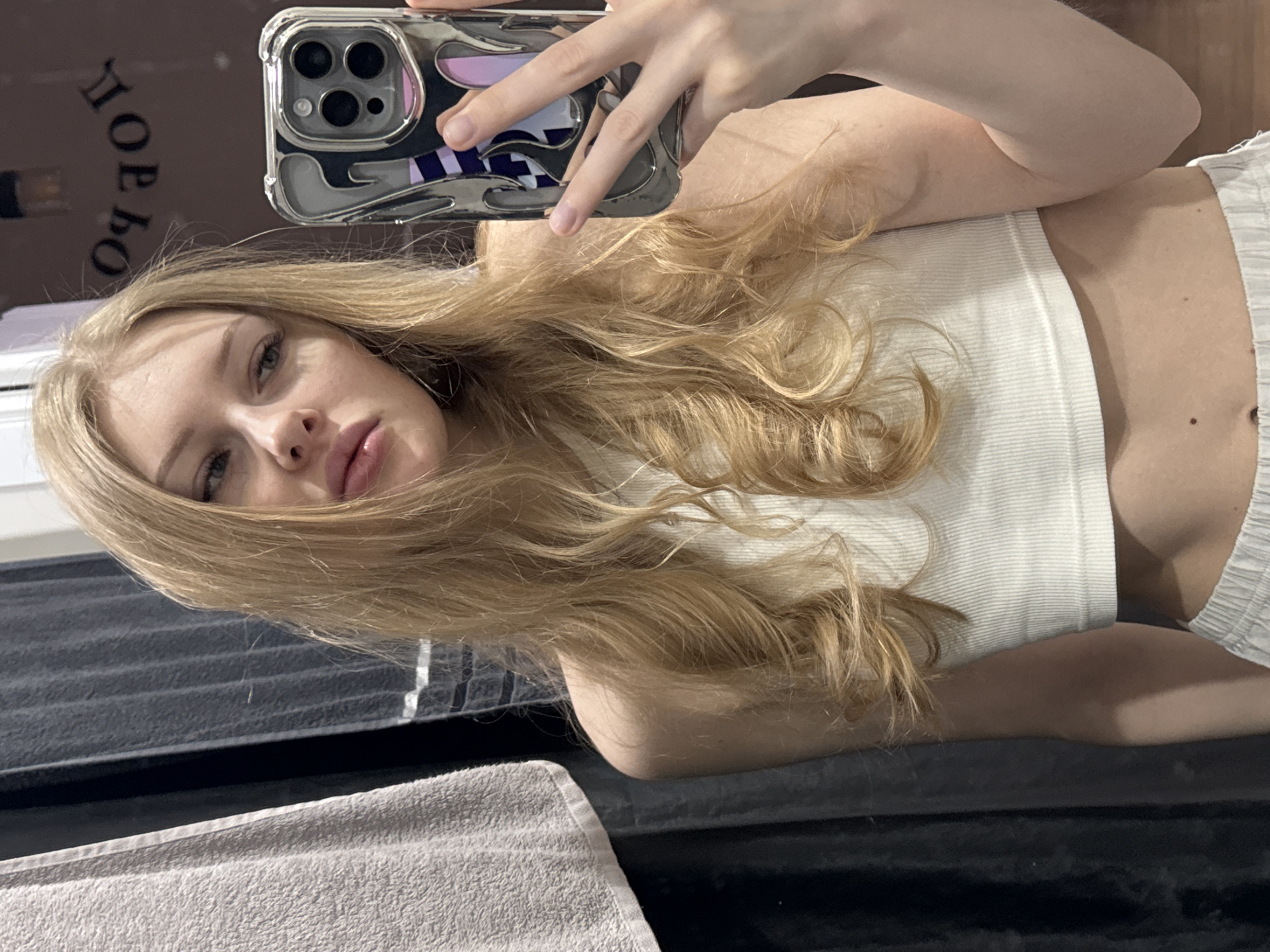 A woman with long blonde hair is taking a selfie using an iPhone with a reflective, decorative case. She is wearing a white ribbed tank top. The background appears to be a dimly lit interior space with dark surfaces. "ЛЕОБО" is reflected, backwards, in the mirror. This word indicates this might be taken at "LEOBO", a chain of beauty salons in Russia. Given the Cyrillic writing and "LEOBO" mentioned, it's possible the location is Perm, Russia, where the chain exists. A light gray towel is visible in the foreground.