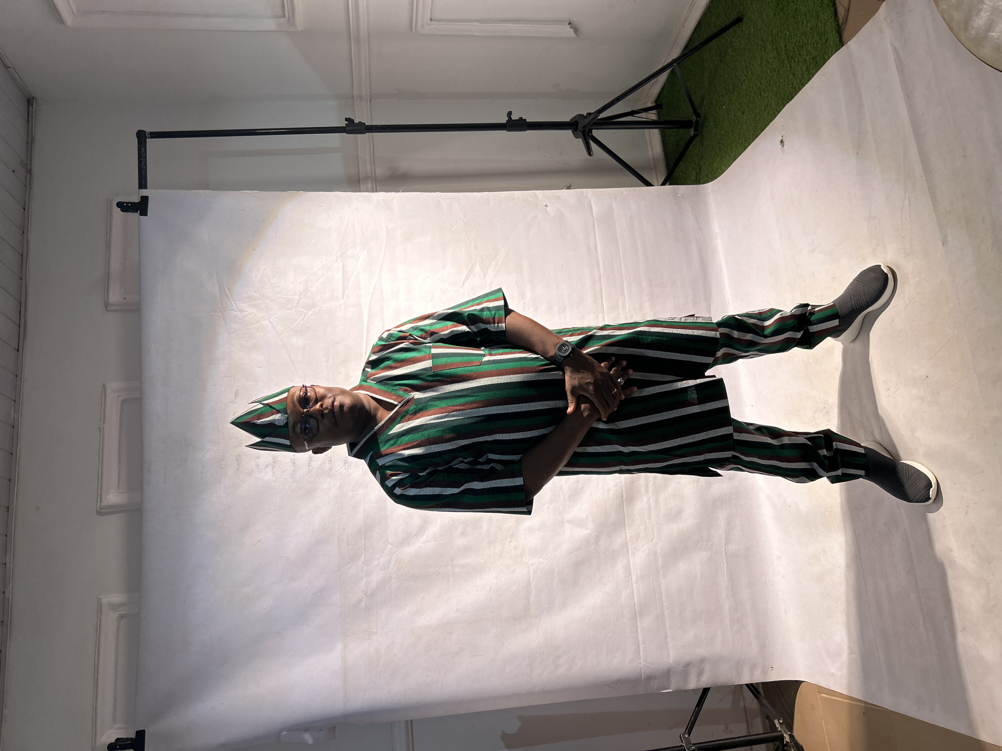 A person wearing a striped green, brown, and white outfit with a matching cap and grey shoes is standing on a white backdrop. The backdrop is set up in a room with white walls and moldings. A black backdrop support stand is visible behind the person and to the left. Artificial turf is partially visible in the upper right corner. The person is wearing glasses and a watch, with their hands clasped. No evidence indicates this photo was taken in Jos, Nigeria.