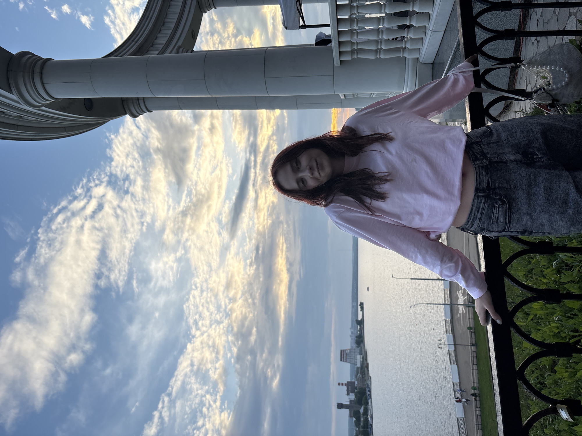A young woman with auburn hair stands on a balcony overlooking a river in Izhevsk, Russia. She is wearing a light pink long-sleeved crop top and dark grey jeans. Her left hand rests on the black railing in front of her. She holds a light-colored beaded handbag with her right hand. In the background, a wide river is visible, with industrial buildings and a city skyline on the opposite bank. The sky is filled with dramatic clouds, illuminated by the setting sun. The architectural style of the balcony suggests a monumental building, possibly a public space or monument.