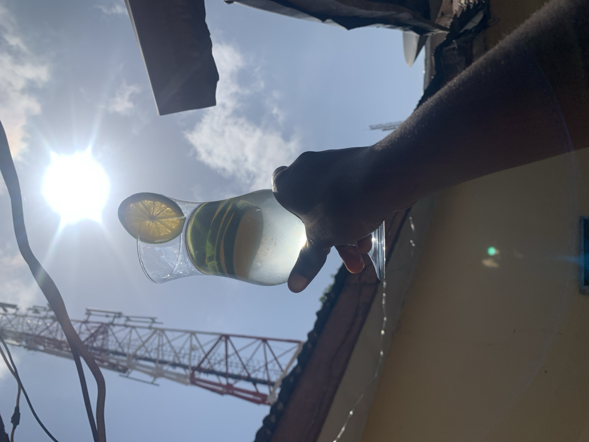 The image depicts a person holding a glass filled with a clear liquid, lemon slices, and cucumber slices. The sun is in the background with a partly cloudy blue sky. A large communications tower is also visible in the background. The photo appears to have been taken from a low angle, looking upwards. The location is Rafin Par, Nigeria. The person is holding the glass up, presumably to capture the bright sky and sun in the background. Water droplets are also visible dripping from the glass.