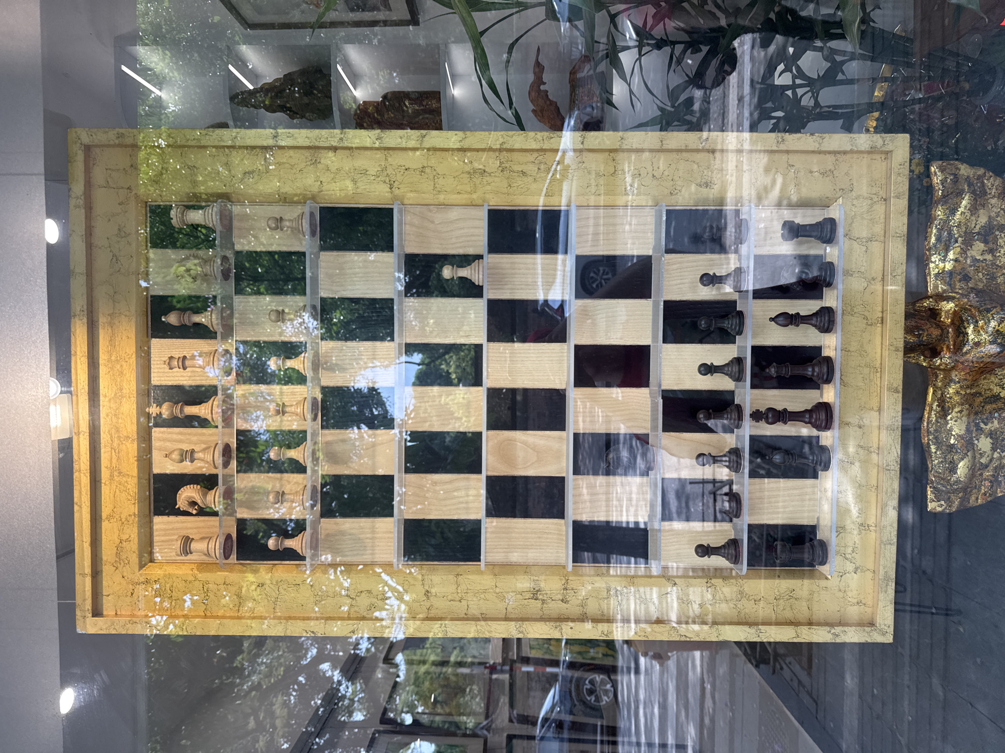 The image shows a chess set displayed behind a glass window. The chessboard is set with wooden pieces, arranged in starting positions. The board has alternating light and dark squares. A gold-colored frame surrounds the chessboard. Reflections of the outside environment, including trees, buildings, and possibly traffic, are visible on the glass.
While the request asks that the location be identified as Hà Nội, Vietnam, there is no explicit detail in the image to confirm its location. The objects present in the photo could conceivably exist in many parts of the world.