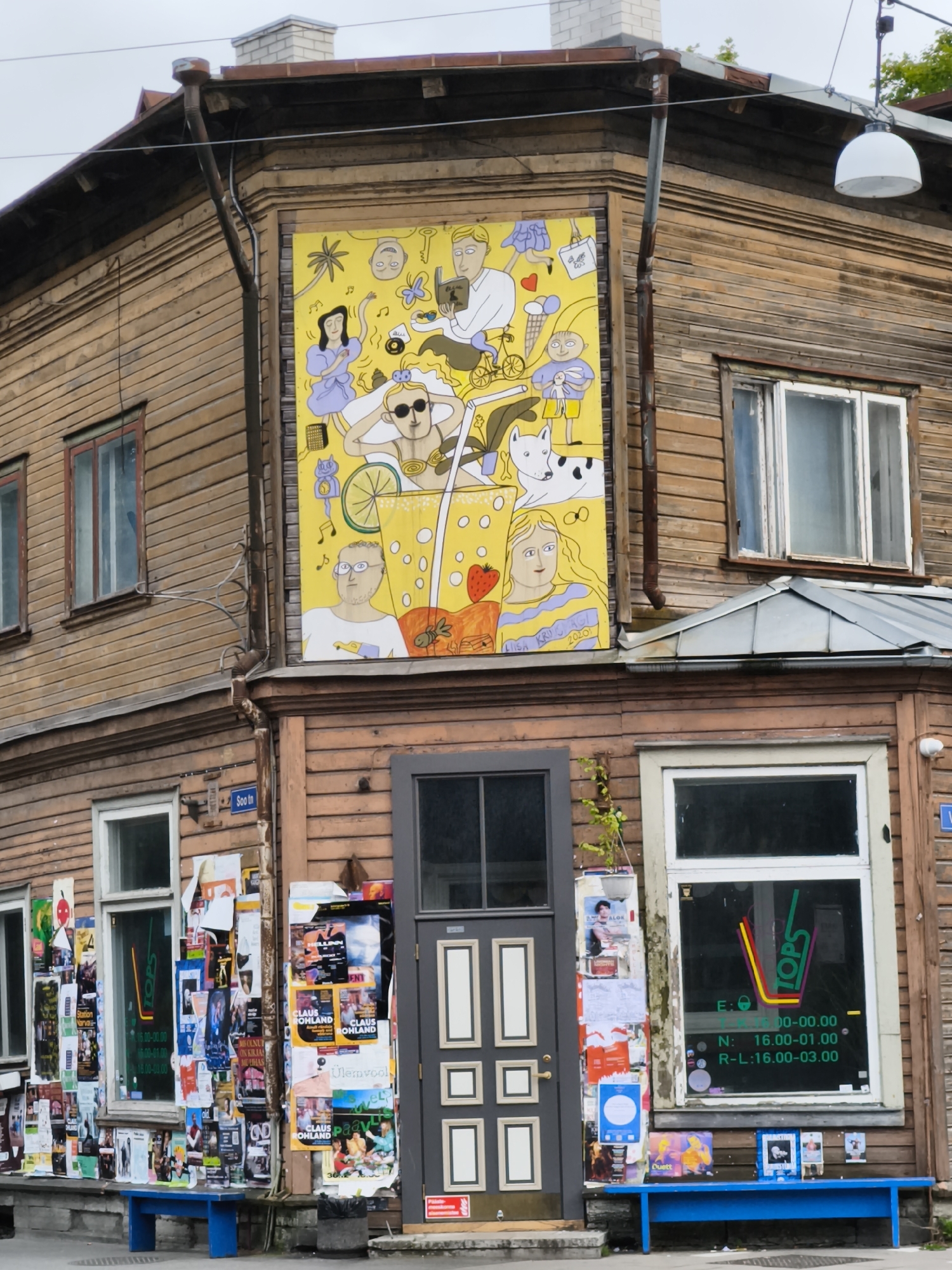 This is a photograph of a wooden building in Tallinn, Estonia, featuring a large mural on its facade. The building has a rustic appearance with weathered wooden planks and multiple windows. The mural, which dominates the upper part of the building, is a colorful and whimsical depiction of people and objects against a bright yellow background. It includes stylized figures, a bicycle, a dog, and various symbolic elements such as hearts and musical notes.
Below the mural, on the ground floor, there is an entrance with a gray door and several windows. One of the windows displays a sign with the text "TOPS" in green and red lettering. The windows and the area around the entrance are adorned with a multitude of posters and flyers, creating a visually cluttered but vibrant effect. A bright blue bench is placed in front of the building, and a trash can is visible next to it. The overall scene suggests an urban environment with a blend of historic architecture and contemporary street art.