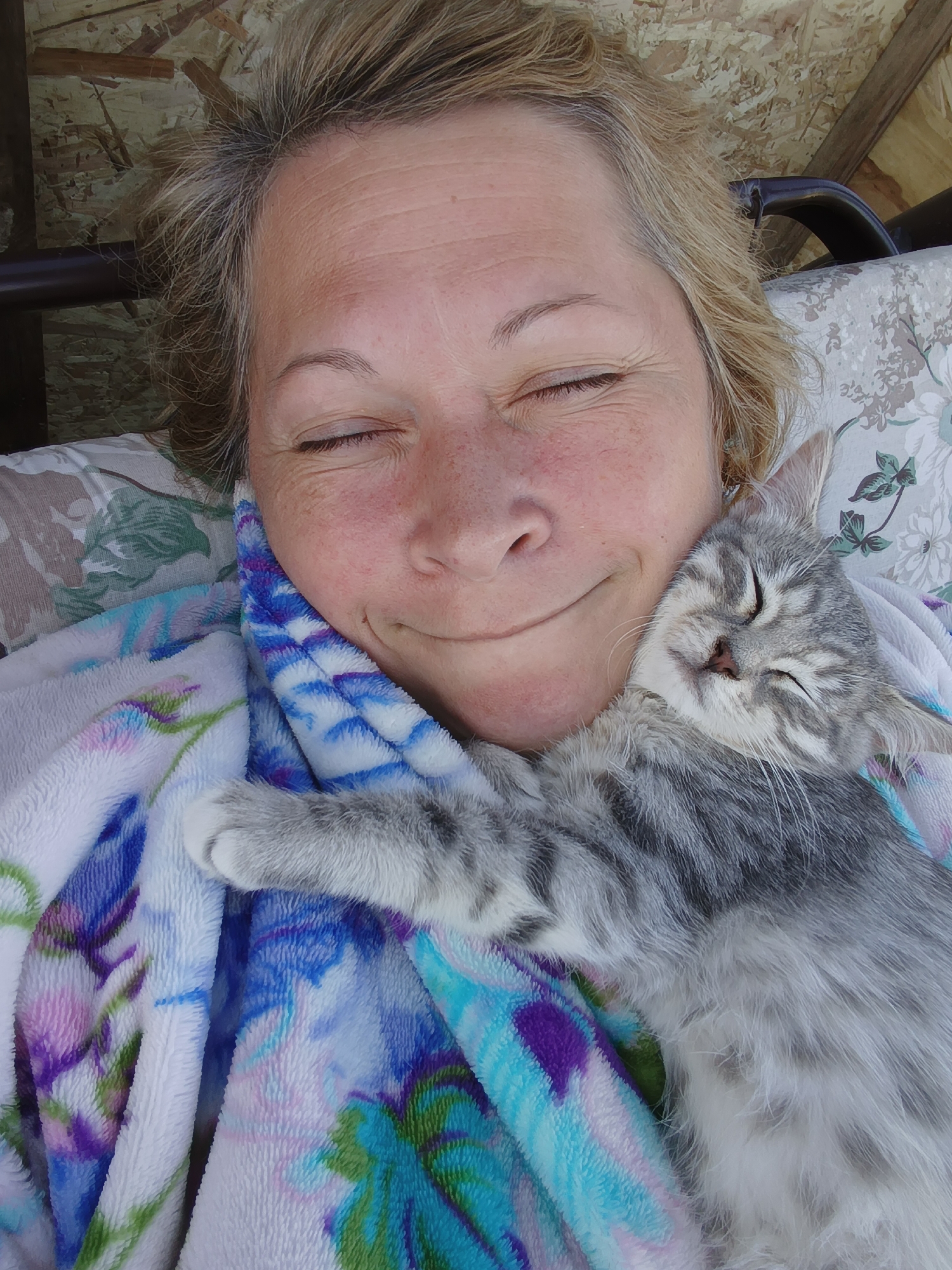 A woman with blonde hair and closed eyes is resting her head against a small, grey tabby kitten, which is also asleep with its eyes closed. They are nestled together under a soft blanket with a floral pattern in shades of blue, purple, and green. The background appears to be an outdoor setting, possibly a porch or patio, with wooden paneling visible at the top and a hint of outdoor furniture on the right. The scene conveys a sense of peace and affection between the woman and the kitten. This image does not provide any information about the location in the city of Ivanovskoe, Russia.