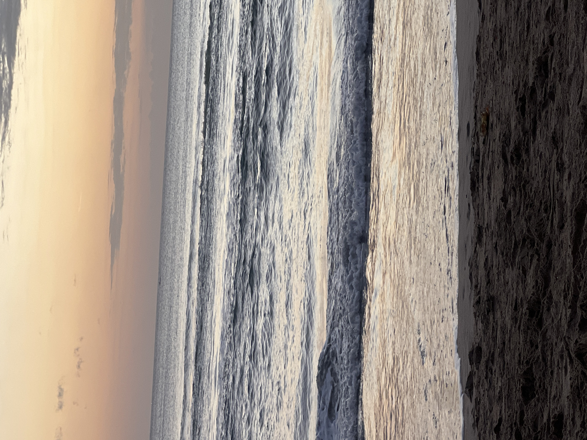 The image depicts a serene beach scene at dusk. The sky is painted with soft hues of orange and yellow, transitioning to a pale pink near the horizon. Wispy clouds are scattered across the sky, adding texture to the twilight canvas. The ocean stretches out to meet the sky, with gentle waves rolling towards the shore. The water reflects the warm colors of the sunset, creating a shimmering effect. In the foreground, dark, wet sand covers the beach, with small ripples and indentations from the receding tide. A solitary piece of debris, possibly a shell or small piece of plastic, is visible on the sand. The overall atmosphere is one of tranquility and natural beauty, characteristic of a beach location in Kuta, Indonesia.