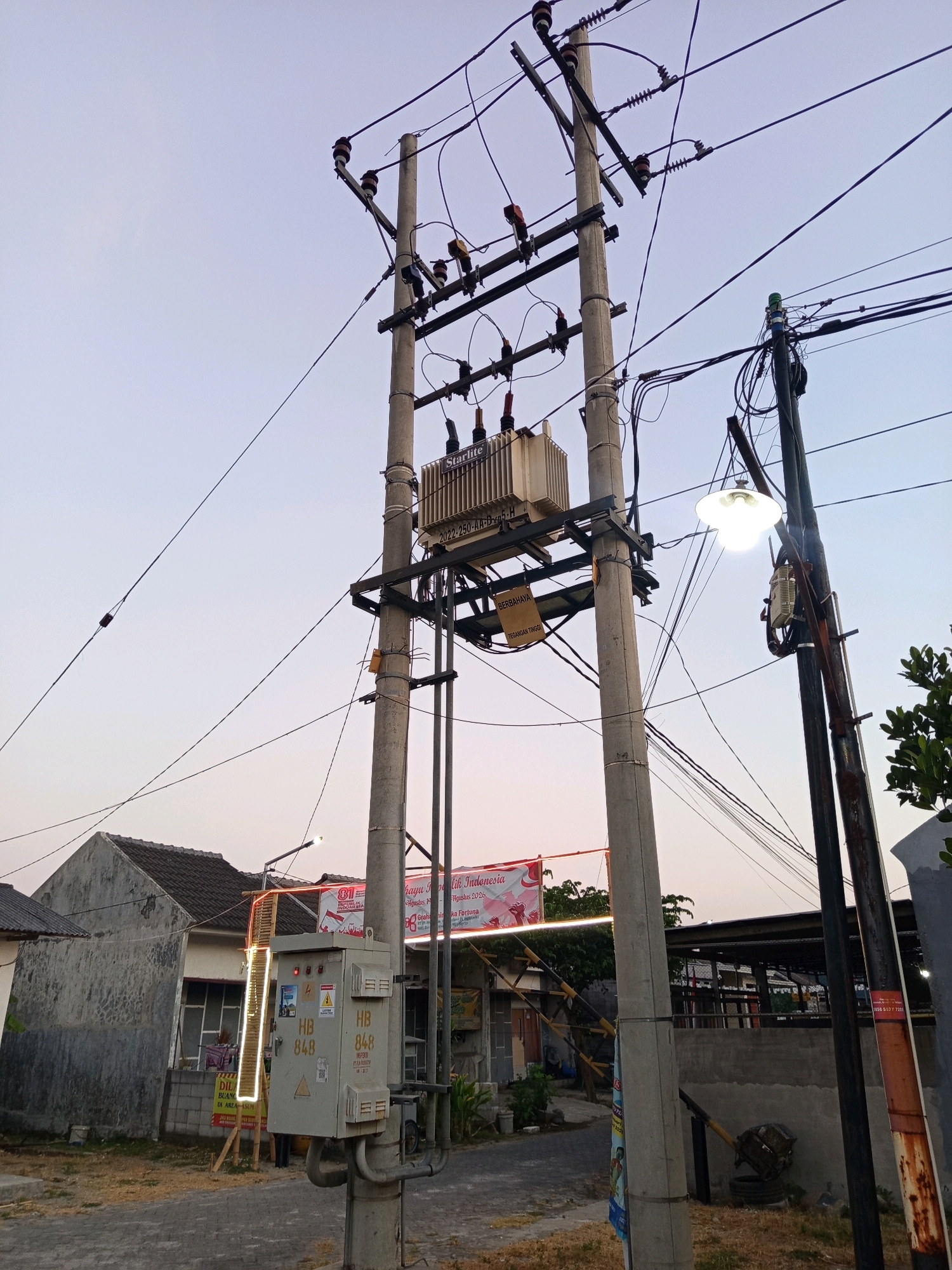 The image depicts an electrical pole with a transformer and a street lamp in Ngastemi, Indonesia. The pole is made of concrete and supports several power lines. A transformer, labeled "Starlite 2022-250-AA-25H," is mounted on the pole. A street lamp with a white, bowl-shaped shade illuminates the scene. In the background, a house with a gray roof and white walls is visible, along with other utility poles and wires. A banner with Indonesian text, possibly related to a national holiday, is also present. A gray electrical box is attached to the base of the main pole, featuring labels such as "HB 848" and "INSPEKSI." The environment appears to be a residential area, with dry grass and a paved road visible. The sky is overcast with a pale orange hue, suggesting either dawn or dusk. No people are clearly visible in the image.