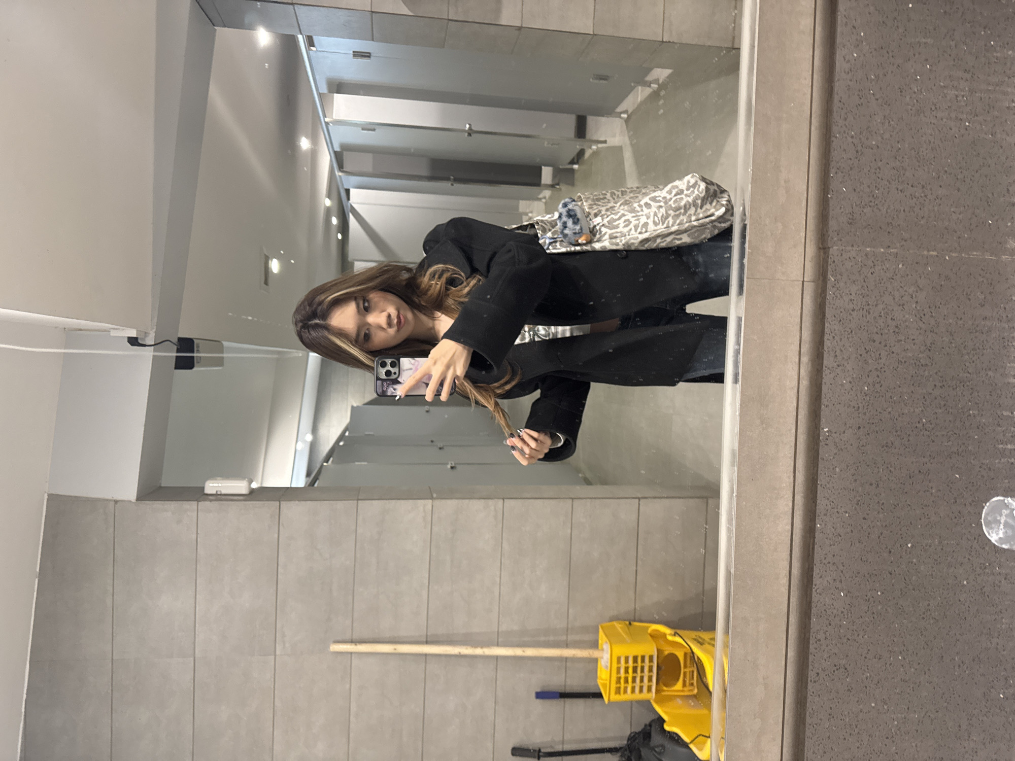 A woman takes a selfie in a public restroom, evidenced by multiple stalls visible in the background mirror. She holds a smartphone in her left hand, her right hand playing with the ends of her hair. She is wearing a black coat, jeans, and a patterned purse hangs over her shoulder. The restroom floor is littered with cleaning supplies, a mop bucket and mop being the most prominent. The wall is tiled in a light grey. There is no contextual information in the image indicating a location in Guazhou County, China.