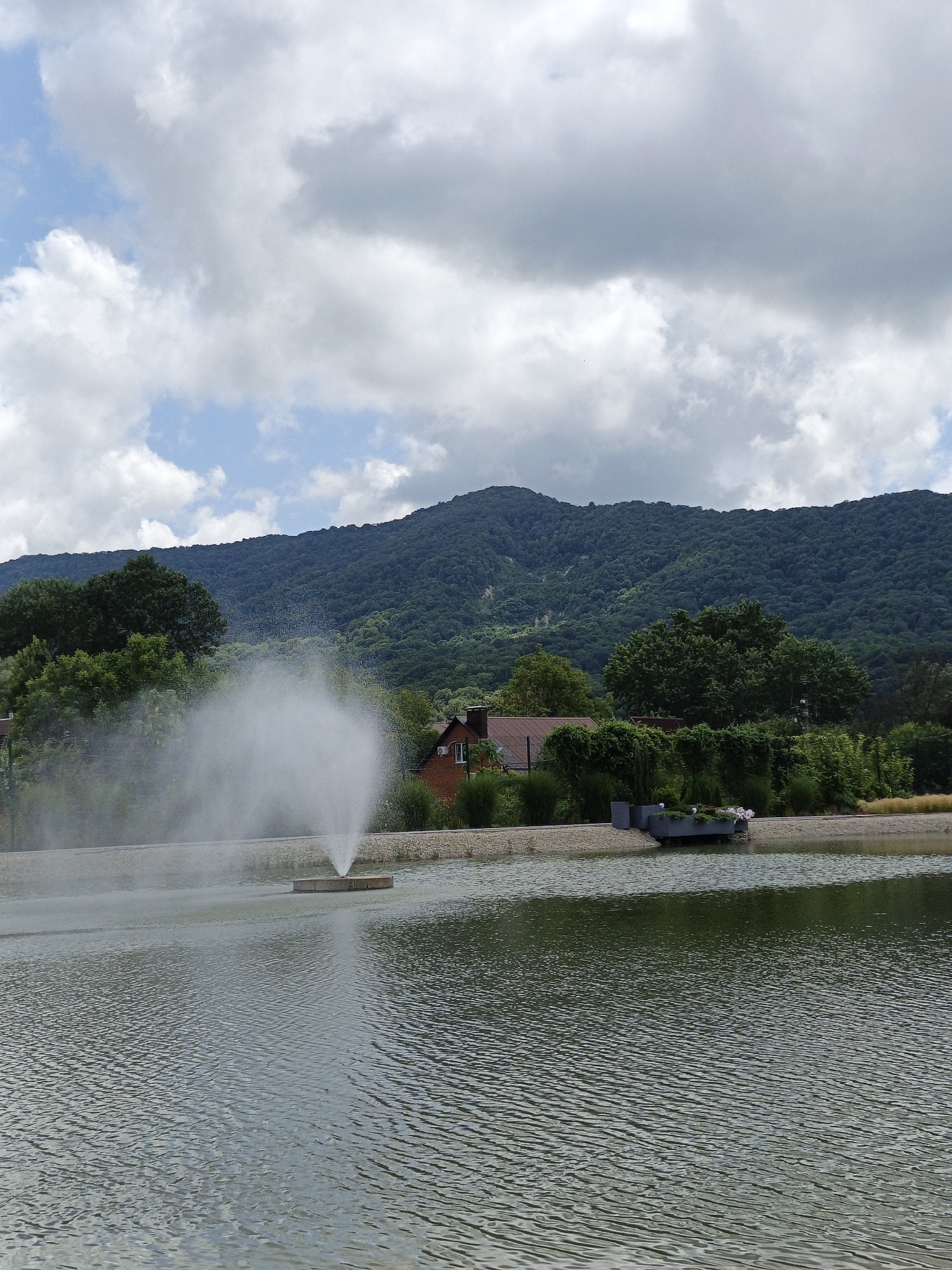 The image depicts a serene landscape scene featuring a body of water with a fountain actively spraying water into the air. In the background, lush green trees form a dense forest that covers the slopes of a large mountain. A red-roofed building, possibly a house, is partially visible among the trees. The sky above is a mix of blue and white clouds. The scene appears to be a natural park or a landscaped area, likely in or near the city of Pshada, Russia, given the presence of mountainous terrain and dense vegetation.