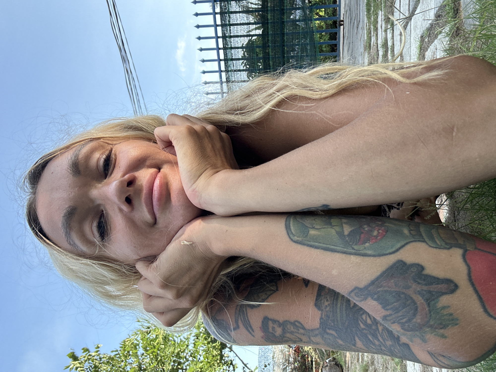 The image shows a close-up, low-angle, selfie-style shot of a woman outdoors, with her face propped up on her hands. She has long, blonde, wavy hair, and her arms are covered in tattoos. In the background, there is a blue sky, some greenery, a fence, and what appears to be a paved walkway with grass growing through it. The scene suggests a casual, outdoor setting, possibly a yard or garden area. The lighting indicates it is daytime. It is not possible to determine the specific location in Mũi Né, Vietnam, from the visual information provided.
