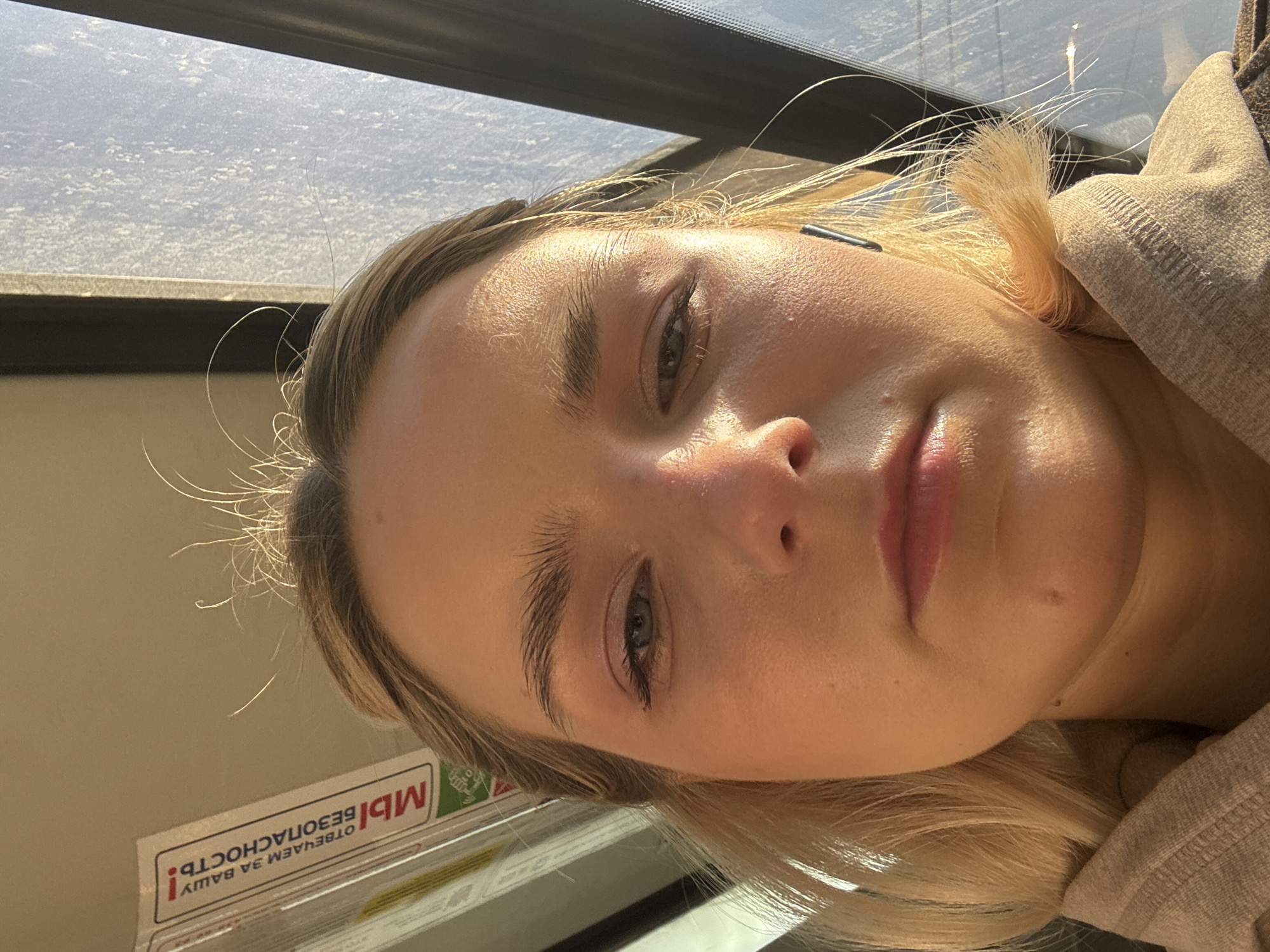 The image features a close-up, rotated portrait of a young woman, likely a selfie. She has fair skin, blonde hair, and light-colored eyes, looking directly at the camera with a neutral expression. Bright natural light, possibly sunlight, illuminates her face from the top right, creating highlights and a slight sheen on her skin. Wisps of her blonde hair catch the light around her face. A dark earbud is visible in her left ear. She is wearing a light-colored, possibly beige or grey, hooded garment.
The setting appears to be the interior of a public transport vehicle, such as a train or bus, during daytime. A window is visible in the upper part of the image, showing a bright, somewhat blurred exterior, possibly a sky with some cloudiness or dust on the glass. Below the window, a light-colored interior wall is present.
A prominent detail is a rectangular sign with Russian text, positioned upside down in the lower left background due to the image's rotation. The text on the sign reads: "МЫ ОТВЕЧАЕМ ЗА ВАШУ БЕЗОПАСНОСТЬ!" which translates to "WE ARE RESPONSIBLE FOR YOUR SAFETY!". This type of sign is commonly found in Russian public transportation.