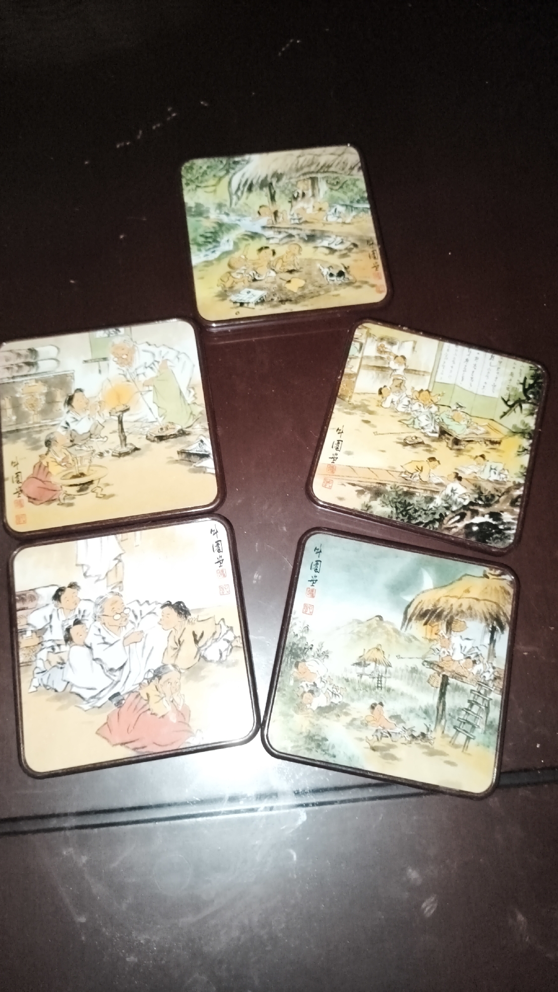 The image displays five square coasters arranged in a fan-like formation on a dark, reflective surface, possibly a wooden table. Each coaster features a colorful, traditional East Asian painting depicting scenes with figures in historical attire. One coaster shows an elderly person with a long beard, seemingly instructing younger individuals around a small fire or brazier. Another depicts a group of people, including children, gathered in what appears to be a courtyard or interior setting with wooden screens. A third coaster illustrates a lively outdoor scene with multiple figures engaged in various activities near thatched-roof structures and foliage, under a crescent moon. The remaining two coasters also showcase similar scenes with groups of people in traditional clothing, some sitting and others standing, amidst natural or architectural elements. The coasters appear to be made of a material with a glossy finish, and the dark background shows some dust or debris. The arrangement of the coasters suggests they are part of a set, possibly for decorative or functional use. The location is described as South Jakarta, Indonesia.