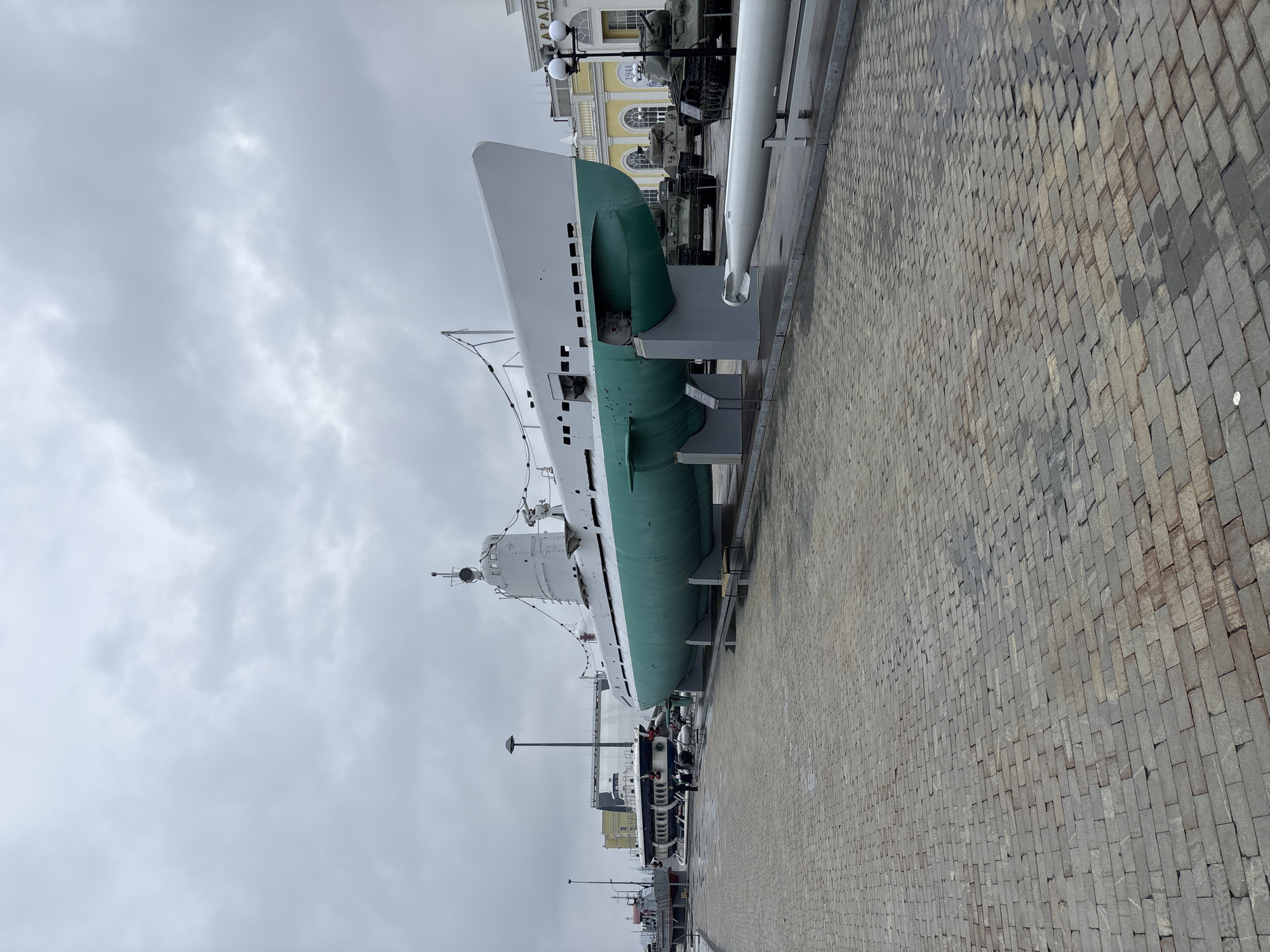 In Verkhnyaya Pyshma, Russia, a museum exhibits a large submarine on land, painted white on top and green on the bottom, supported by grey stands. To the right of the submarine, a white torpedo is displayed horizontally on its side. Behind the submarine, several military tanks are parked on a paved area. In the background, a light-yellow building with arched windows and columns is visible, with the word "ПАРАД" (Parade) emblazoned on it. The sky is overcast with grey clouds. The foreground consists of a brick-paved walkway with some wet patches, indicating recent rain. Several lampposts are present along the walkway.