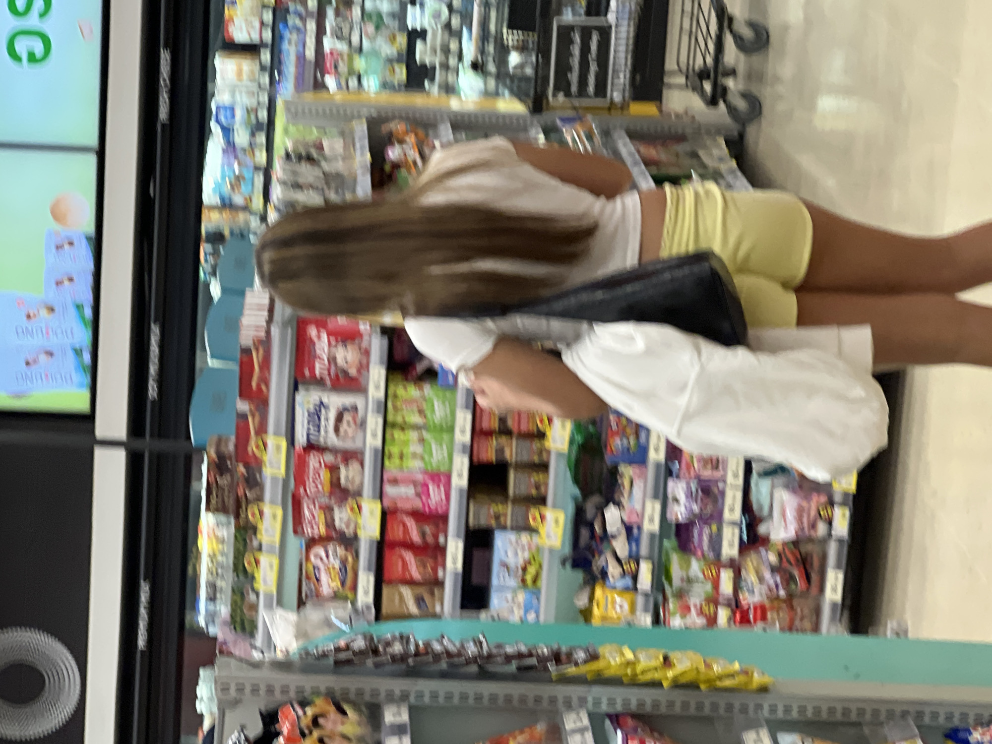 The image depicts the interior of a convenience store, possibly a 7-Eleven, in Baan Chaweng, Thailand. A young woman with long brown hair, wearing a white t-shirt and yellow shorts, is walking away from the camera, carrying a white bag and a dark handbag. She is positioned in an aisle with shelves stocked with various packaged goods, including snacks and beverages. The shelves are well-lit, and the floor is a light-colored tile. In the background, other aisles and a shopping cart are visible. The overall environment suggests a typical retail setting.