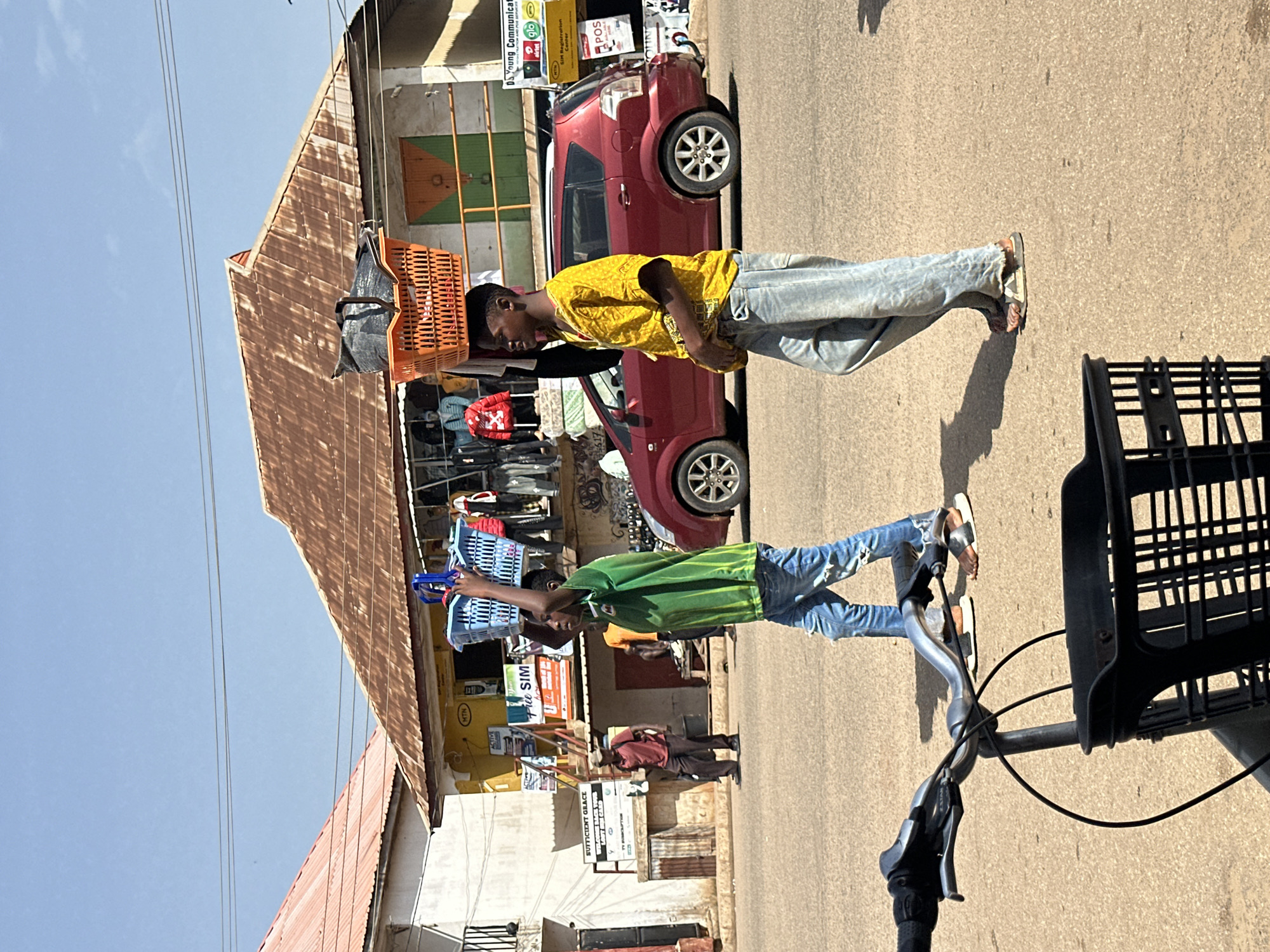The image shows two young men standing on a dusty street in a market area in Bukuru, Nigeria. The scene is bathed in bright sunlight, suggesting it is daytime, likely mid-morning or afternoon, with clear skies and warm weather.
The man in the foreground, wearing a green t-shirt and ripped blue jeans, is carrying two blue plastic baskets. He is looking slightly down and to his right. To his left, further down the street, stands another young man wearing a yellow t-shirt and light blue jeans, also carrying a basket balanced on his head. He appears to be looking down as well.
Behind the first man, a red car is parked on the side of the road. Several stalls or shops are visible along the street, with awnings and displays of goods, typical of a market setting. Some of the goods appear to be clothing, with racks of garments visible. Electrical wires crisscross the sky above the street.
On the buildings in the background, several signs with text are visible, indicating various businesses. Some of the legible text includes "SUFFICIENT GRACE," "MTN," "airtel," and "glo," suggesting a telecommunications and general services area. The presence of SIM registration centers and POS terminals on the signs further contextualizes the commercial activity. The overall impression is of a bustling, everyday scene in a Nigerian market.