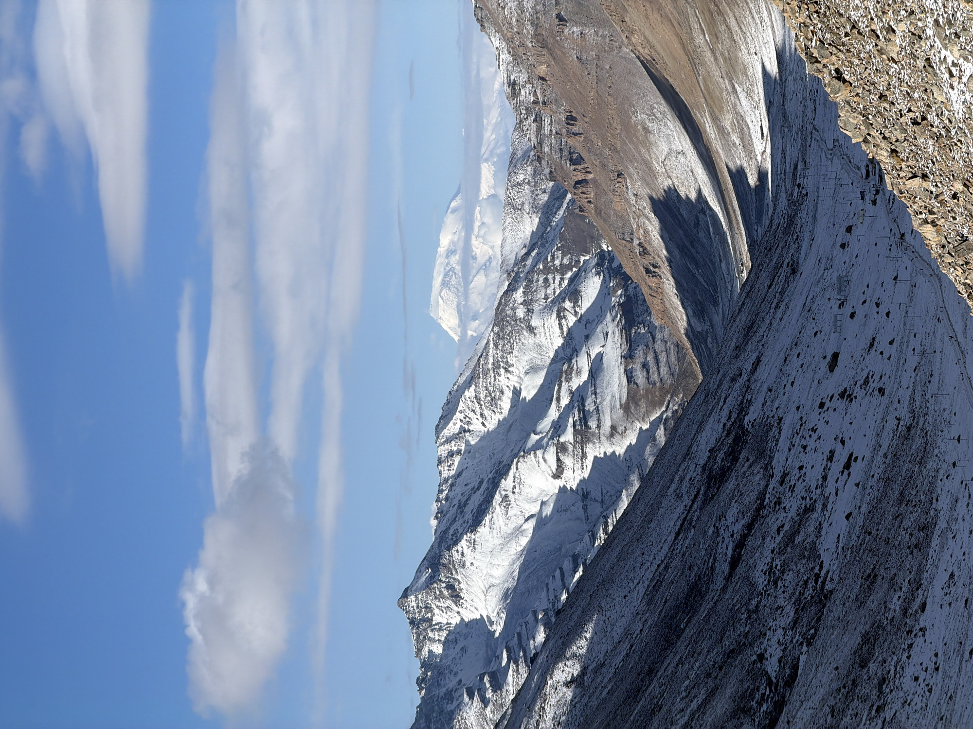 The image presents a majestic high-altitude mountainous landscape under a clear, bright blue sky dotted with elongated white clouds. The scene is characterized by rugged, snow-covered peaks and steep rock faces, suggesting a cold, alpine environment.
In the foreground and extending along the right side, a massive, dark, rocky mountain slope dominates, partially dusted with snow and scree. This slope is illuminated by direct sunlight, casting prominent shadows that emphasize its coarse texture and steep incline. Faint, dark linear structures, possibly indicating telecommunication towers or elements of a ski lift, are sparsely visible on this vast slope.
Towards the center and left, a series of towering snow-capped mountains emerge. Their sharp ridges and deep valleys are defined by a striking interplay of light and shadow; sun-drenched snow gleams brightly, contrasting sharply with the dark, shaded crevices and rock faces. Further into the distance, additional layers of hazy, snow-dusted peaks fade into the atmospheric blue, extending the grandeur of the mountain range.
The sky, occupying the upper left quadrant, is a vivid blue, streaked with delicate, wispy white clouds, typical of cirrus formations at high altitudes. The strong directional shadows imply that the photograph was taken during either the morning or late afternoon.
No people or explicit activities are depicted in the scene. There is no visible text. The overall impression is one of immense natural power, solitude, and the breathtaking scale of a mountainous region in China.