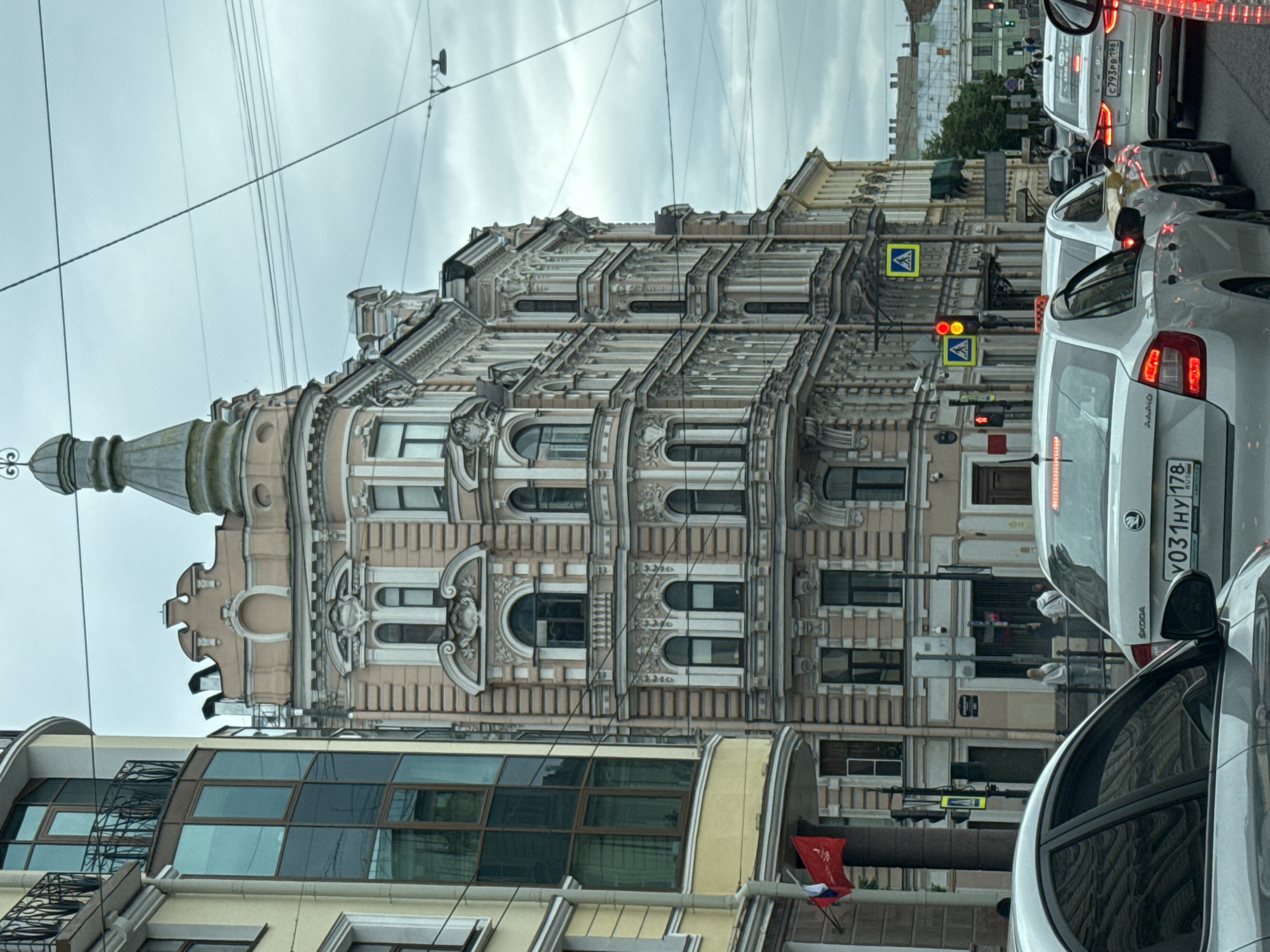This is a medium-shot, street-level, eye-level, slightly angled photograph taken in Saint Petersburg, Russia, on a cloudy day. The primary subject is a multi-story, ornate building with light pink and beige facades, featuring numerous windows, decorative moldings, and a distinctive domed structure at the top. To the left, a modern building with a curved glass facade and a black metal balcony is visible. The street is congested with traffic, including several white sedans and what appears to be a taxi. A red flag with a white and blue emblem is displayed on a pole attached to the modern building. Pedestrian crossings and traffic lights are present, with some people visible on the sidewalk in the background. The overall atmosphere is urban and slightly overcast.