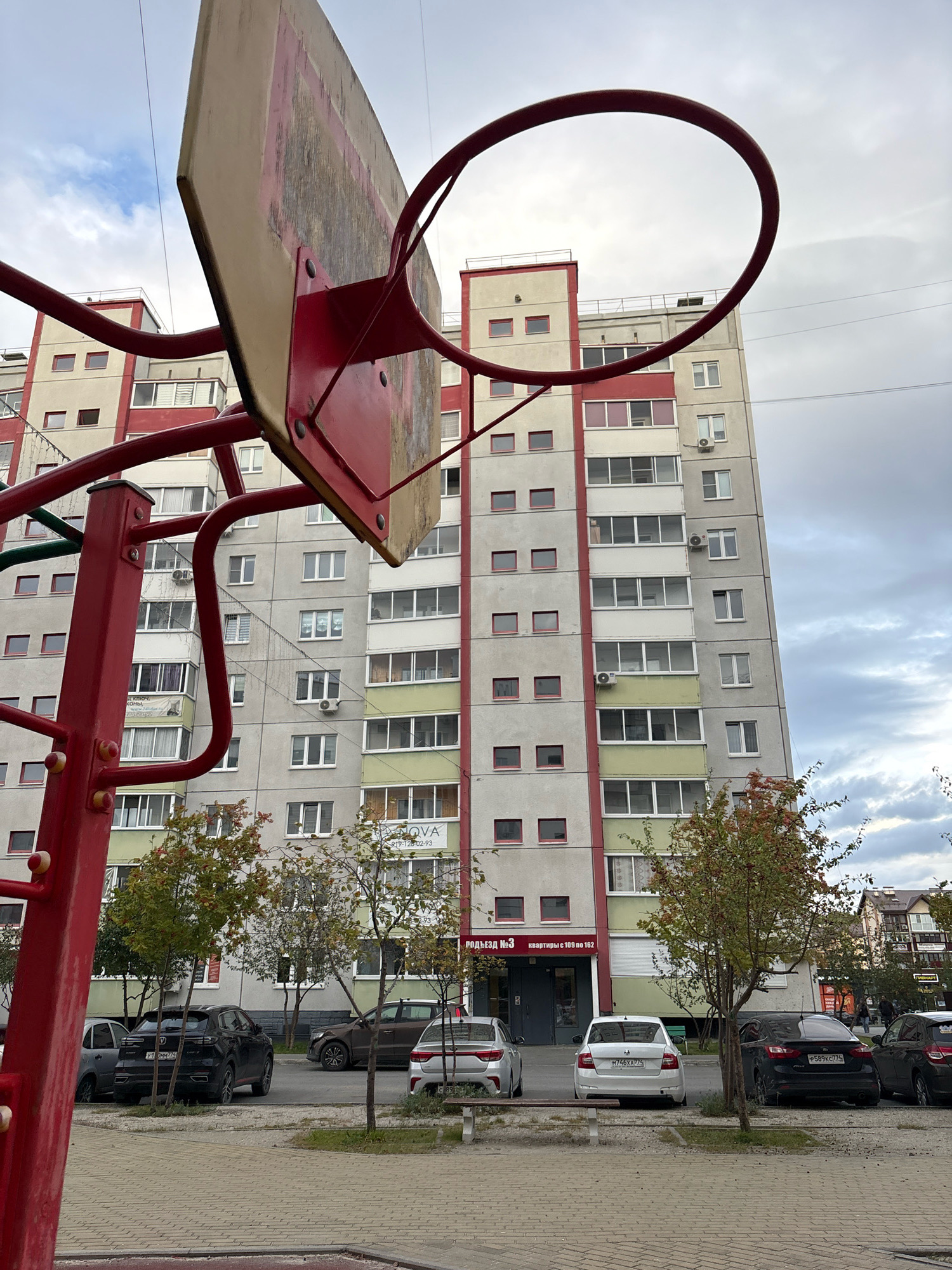 In the city of Zapadny, Russia, a residential building stands tall under a cloudy sky. The building, characterized by its grey facade with vertical red and light green accents, features numerous windows and balconies. In the foreground, a red basketball hoop and backboard are partially visible, suggesting the presence of a playground or sports facility. Several cars are parked on the asphalt in front of the building, with trees and vegetation interspersed between the parking area and the building. The scene is one of everyday urban life in a residential neighborhood.