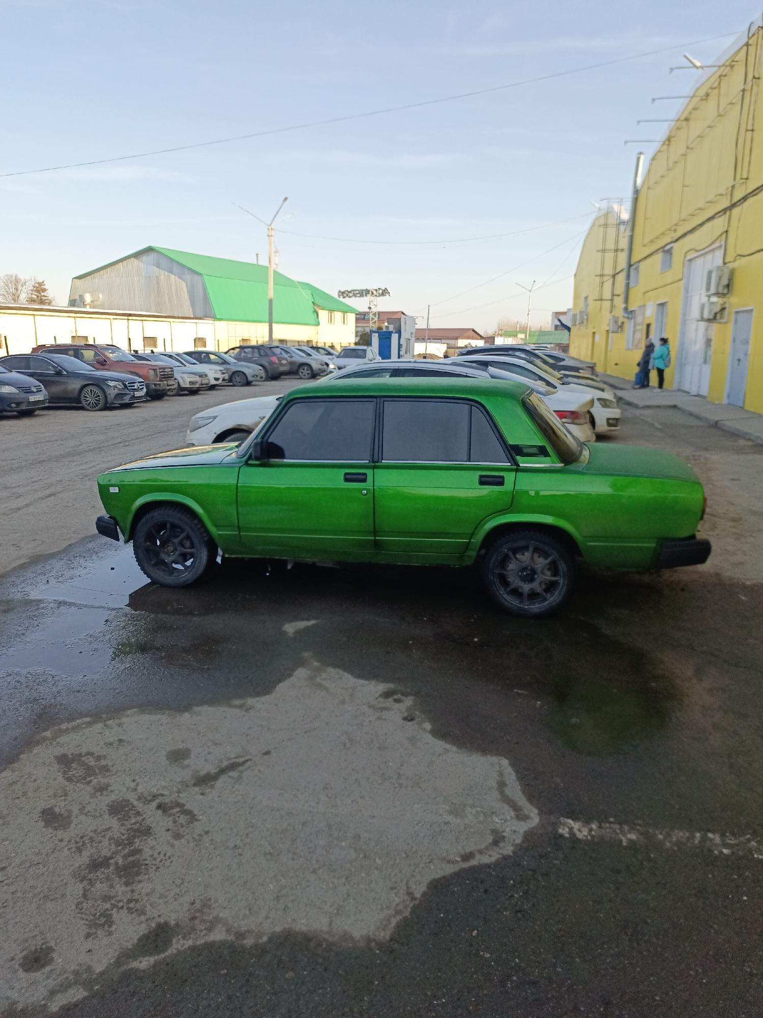 A green Lada sedan sits in a parking lot in Chaikovsky, Russia. The parking lot is wet and stained. Other cars are parked nearby. Behind the cars is a building with a green roof and a yellow building. Two people stand on the sidewalk in front of the yellow building. The sky is blue with some clouds.