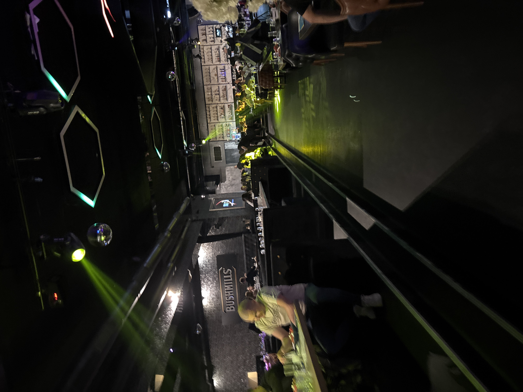 This image captures the interior of a dimly lit, contemporary bar or restaurant located in Sofia, Bulgaria. The black ceiling features exposed metallic piping, multiple small reflective disco balls, and professional lighting fixtures, including a prominent green spotlight casting a beam towards the left side of the room. Several hexagonal LED light panels with a cyan-green glow are also integrated into the ceiling.
The left wall is covered with dark, textured wallpaper and displays a large, backlit "BUSHMILLS" sign. Seating arrangements include dark upholstered booths and tables situated on both a main floor level and an elevated platform to the left. In the foreground, a fair-skinned male with short, light-colored hair, wearing a light-colored t-shirt with a "GUESS" logo and dark pants, is seated at a table on the raised platform. He is observed looking down at a plate of food, possibly a salad, and several glasses.
Towards the rear and right, a brightly illuminated bar area is visible, showcasing numerous bottles arranged on shelves against a wall. Multiple patrons are seated at tables and booths throughout the venue, appearing to be dining and socializing. The dark floor reflects some of the green light emanating from the overhead fixtures. The scene conveys a modern lounge atmosphere with elements characteristic of a nightlife or entertainment venue.