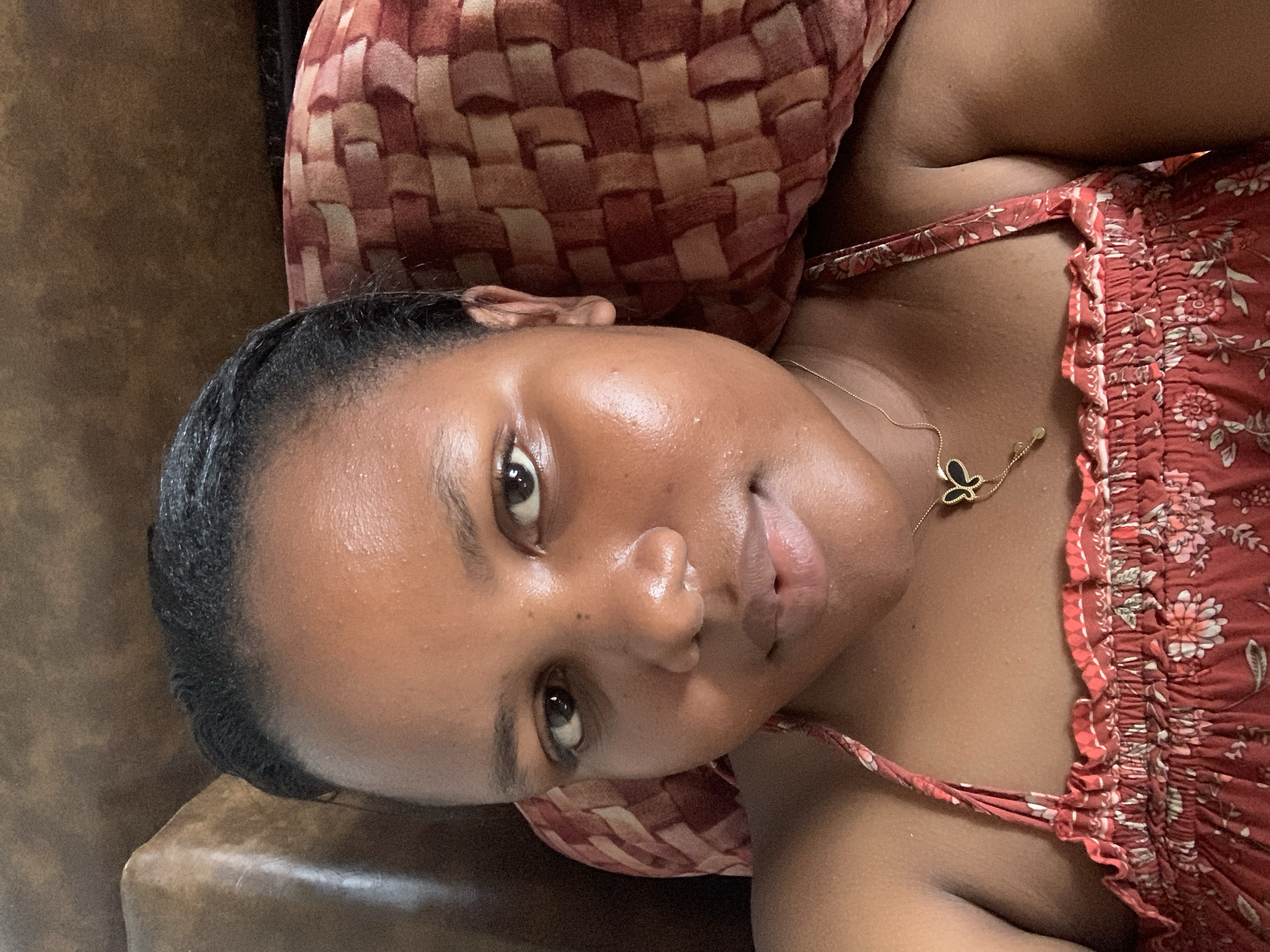 A young woman of color lies on a brown leather sofa, her head resting on a woven, red-and-beige pillow. She wears a floral-patterned, red sundress with thin straps and a gold butterfly pendant necklace. Her hair is pulled back from her face. The location is unknown and cannot be determined from the image.