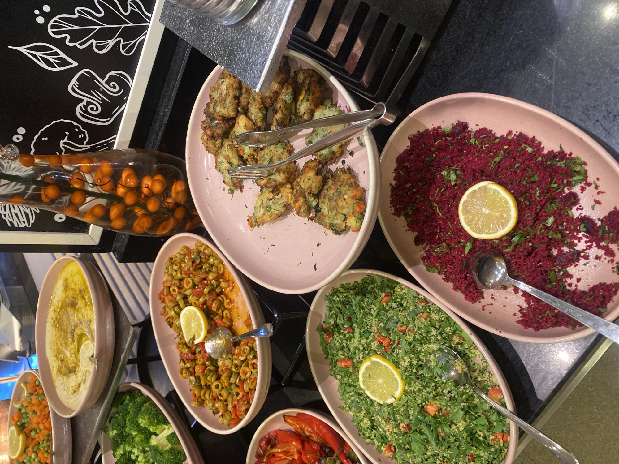 This image captures a buffet spread at a restaurant in Aksu, Turkey. The food items are arranged in large oval-shaped pink platters. In the foreground, there are two dishes side-by-side: one is a vibrant green salad, likely tabbouleh, garnished with chopped parsley and diced red bell peppers, topped with a lemon slice. To its right is a dish of finely grated beetroot salad, also sprinkled with herbs and topped with a lemon slice. A serving spoon is visible in each of these dishes.
Behind these, towards the left, is a platter filled with mixed olives and chopped tomatoes, also adorned with a lemon slice and a serving spoon. Next to this, on the left side of the image, is a bowl containing bright green broccoli florets.
Further back and slightly to the left, there's a large shallow bowl of light-colored dip, possibly hummus, drizzled with olive oil and garnished with a lemon wedge. This bowl is placed on a tiered metal stand. In the upper left corner, a dish with cooked carrots and peas is partially visible.
To the right of the hummus, a platter of golden-brown fritters or patties, likely made with vegetables, is presented with serving tongs. In the background, a clear glass vase contains a stem with small, orange, round fruits, possibly cherry tomatoes or decorative elements, and there's a black board with white decorative drawings of leaves and plants. The overall environment suggests an indoor setting, possibly a hotel or restaurant buffet, with a focus on fresh, colorful dishes. There are no people visible in the frame.
