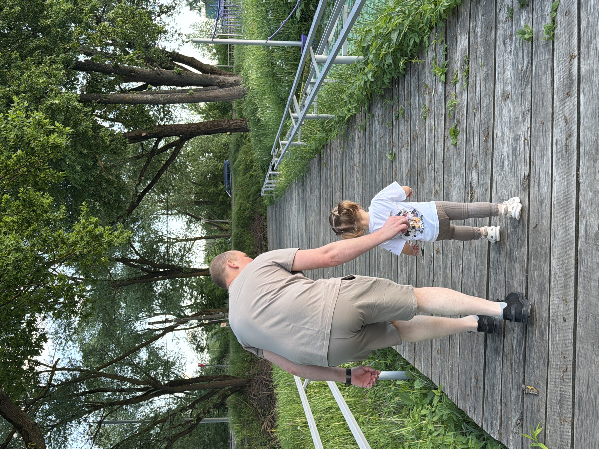 A person and a child stand on a wooden walkway at the park in Sin'kovo, Russia. The person is wearing a light brown t-shirt and shorts with black shoes. The child is wearing a white t-shirt with a design on it and brown leggings. They are next to a green grassy area with trees, and a silver railing borders the wooden walkway.