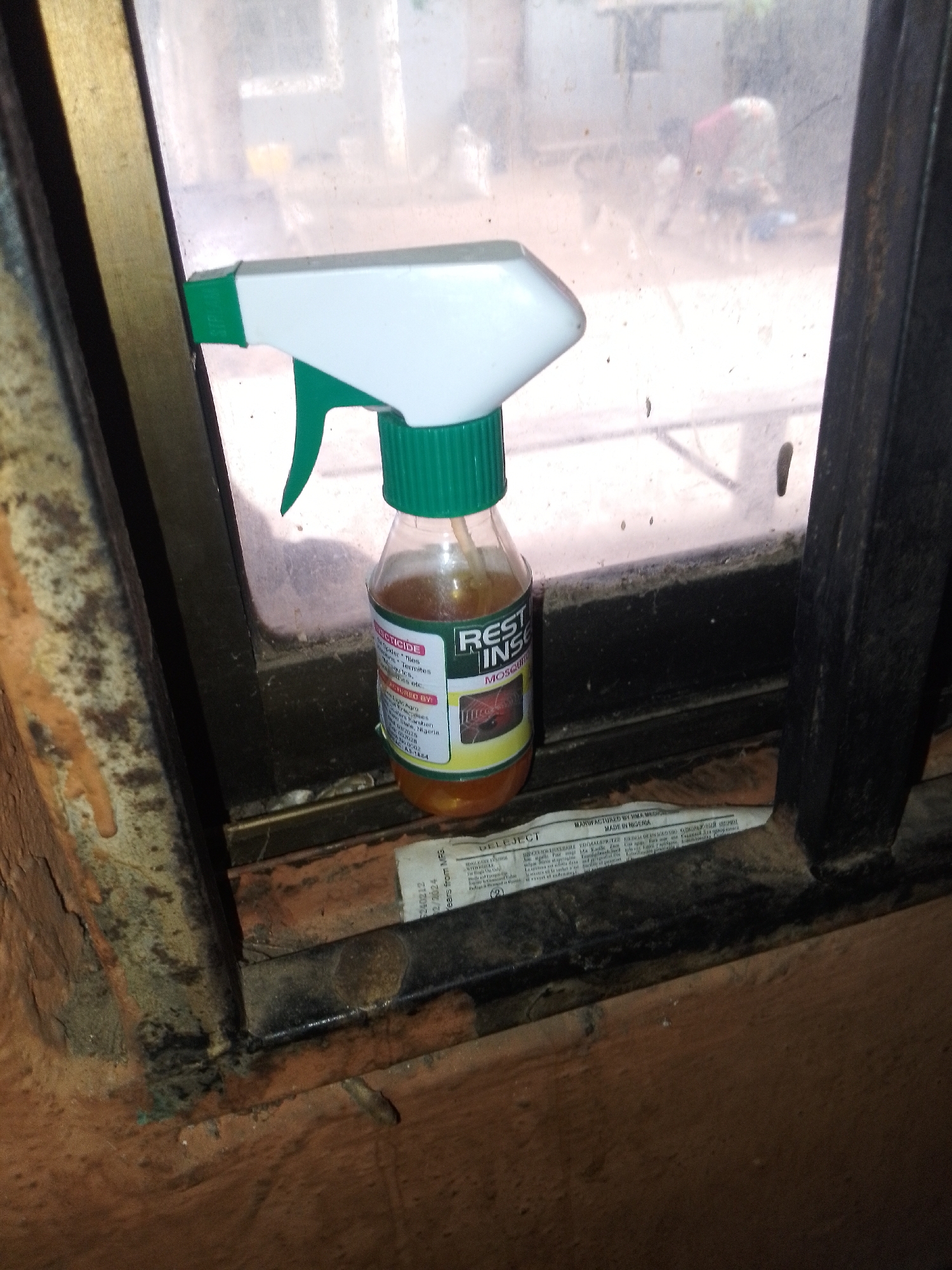 A transparent spray bottle with a white trigger and green nozzle is positioned on a window ledge in Serti, Nigeria. The bottle contains a yellow liquid. A label on the bottle reads "REST INSECT MOSQUITO". The spray bottle is resting on what appears to be a newspaper or pamphlet. The background shows a blurred outdoor scene with a figure and what might be animals. The window frame is dark and appears to be made of metal. The area around the window ledge is dirty and appears to be an interior space.