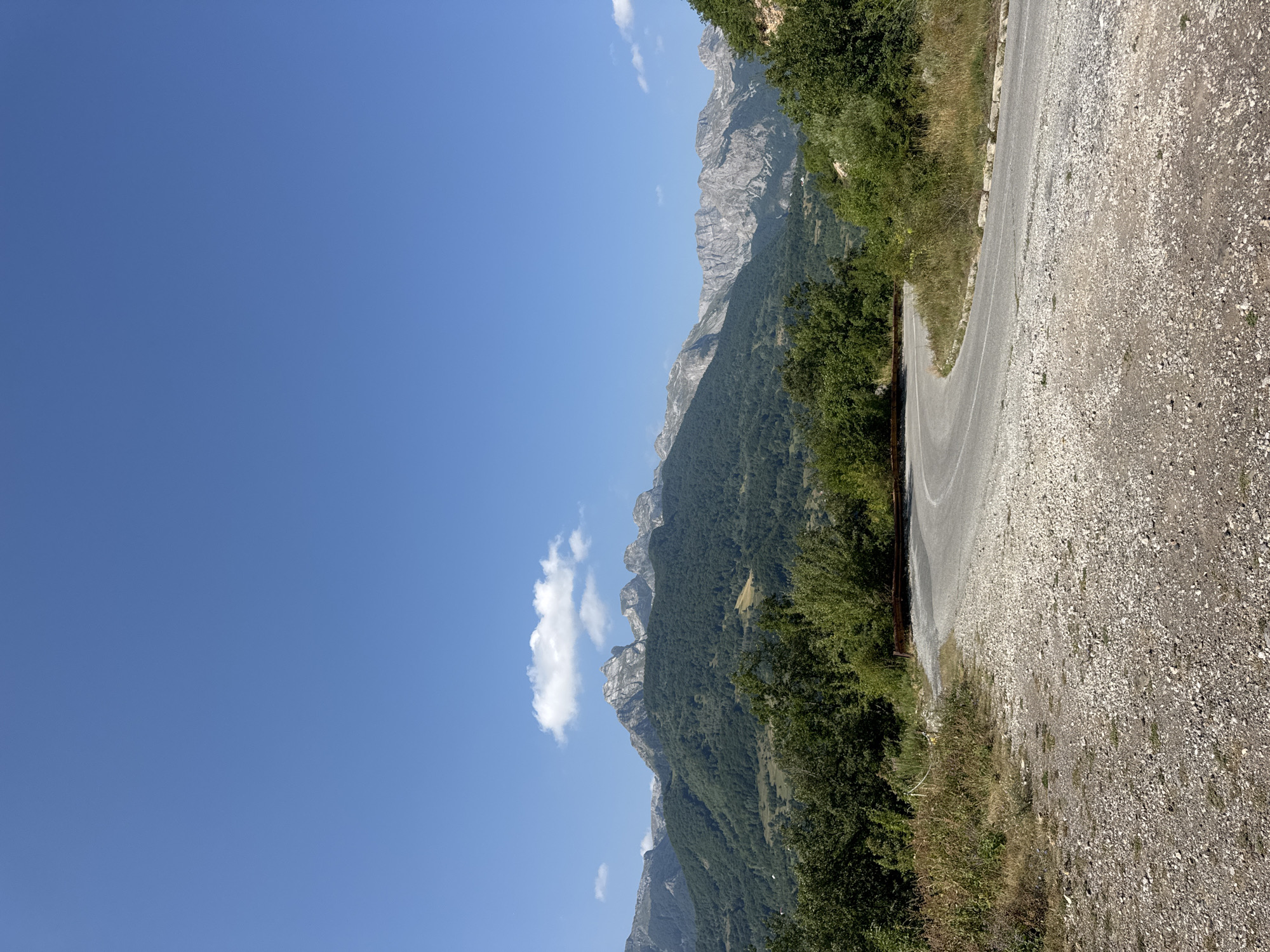 A winding asphalt road curves through a mountainous, forested landscape under a clear blue sky with a single, small cloud. The road, surfaced with gravel on its edges, leads the viewer's eye toward a range of rocky, grey mountains in the distance. These mountains are partly covered by dark green, dense forest. The foreground features dry grass and shrubs along the roadside. The scene depicts a natural, undeveloped area. The image is a vertical shot with the sky occupying the top half and the road and mountains filling the bottom half.