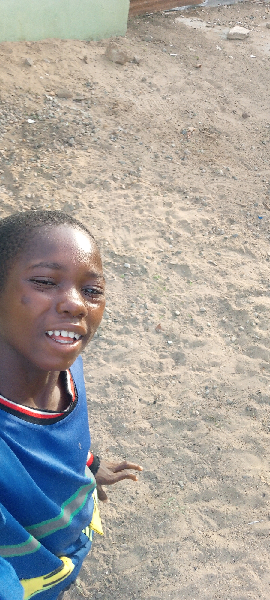 A young, dark-skinned individual is smiling at the camera. They are wearing a blue shirt with red, white, and black trim around the neck and what appears to be a striped design on the sleeve with yellow accents. The background consists of a sandy or dirt-covered ground with scattered rocks and gravel. A light green structure is partially visible in the upper left corner. Given the prompt's information, this scene is potentially located in the city of Adankolo, Nigeria. The photograph appears to be a selfie or taken at close range. The environment suggests an outdoor area with a somewhat undeveloped or construction-related setting.