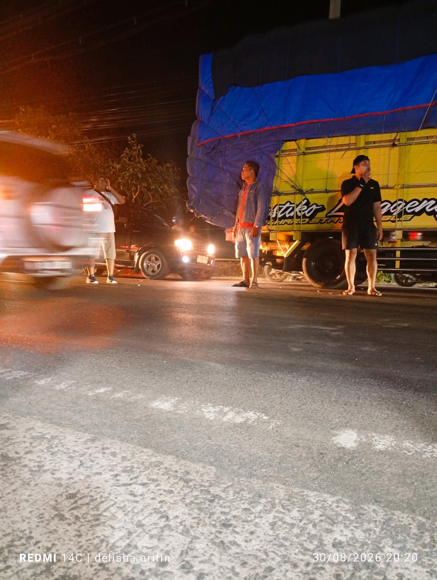 The image depicts a street scene at night in Gambir Kuning, Indonesia. In the foreground, the asphalt road surface is visible, marked with white paint. Two men are standing on the right side of the road. The man on the left wears a denim jacket over a red shirt and light-colored shorts. The man on the right wears a black polo shirt and black shorts. Behind them, a large truck with a yellow body and a blue tarp is parked. The truck has a logo that appears to read "Astiko Langgeng." To the left of the truck, a dark-colored car with its headlights on is parked. Another vehicle, possibly a car, is partially visible on the far left, appearing blurred due to motion or low light conditions, with its headlights illuminating the scene. Trees are visible in the background. The overall lighting suggests it is nighttime, with artificial lights from vehicles and possibly streetlights illuminating the area. The timestamp in the lower right corner indicates the photo was taken on 30/08/2026 at 20:20. The lower left corner contains the text "REDMI 14C | delisha arifin."