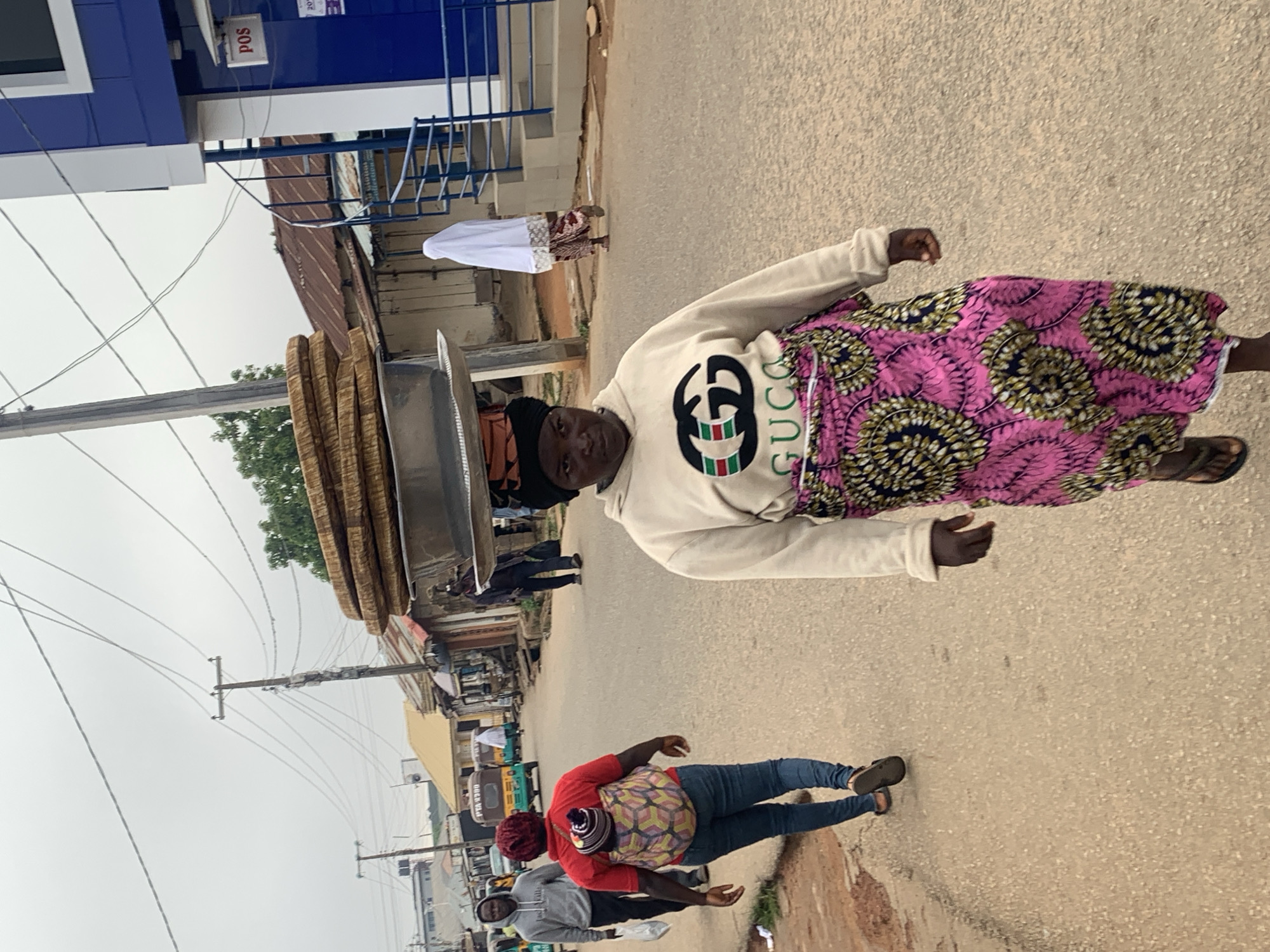 The image depicts a street scene in Jos, Nigeria. The primary subject is a woman in the foreground, facing the camera and wearing a white sweatshirt with a black and green Gucci logo, and a colorful pink and black patterned skirt. She is standing next to a large metal basin filled with stacked, circular food items, possibly bread or pastries.
In the background, several other people are visible, including a person in a white garment walking away from the camera, and another person in a red shirt and jeans on the right. The setting appears to be a bustling street with buildings, utility poles, and overhead wires. The pavement is made of asphalt.
The image appears to be taken during daylight hours, with an overcast sky suggesting potential for rain or a generally cloudy day. The overall atmosphere is that of an everyday street scene in a commercial area. No specific emotions are conveyed by the individuals, who seem to be engaged in their daily activities.
Visible text includes "pos" on a sign attached to a blue building in the upper left corner. The overall orientation of the image is vertical, suggesting it was taken with a phone camera held in portrait mode and later rotated.