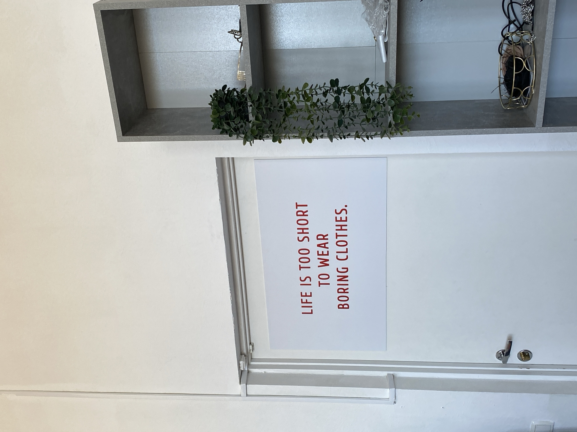 The image shows a close-up of a white door with a sign attached. The sign is rectangular and features a quote in red lettering: "LIFE IS TOO SHORT TO WEAR BORING CLOTHES." Above the door and sign, there is a gray shelving unit with greenery and accessories. The scene is likely located indoors, with plain white walls. The image does not contain people or specific information that would locate it in Izhevsk, Russia.