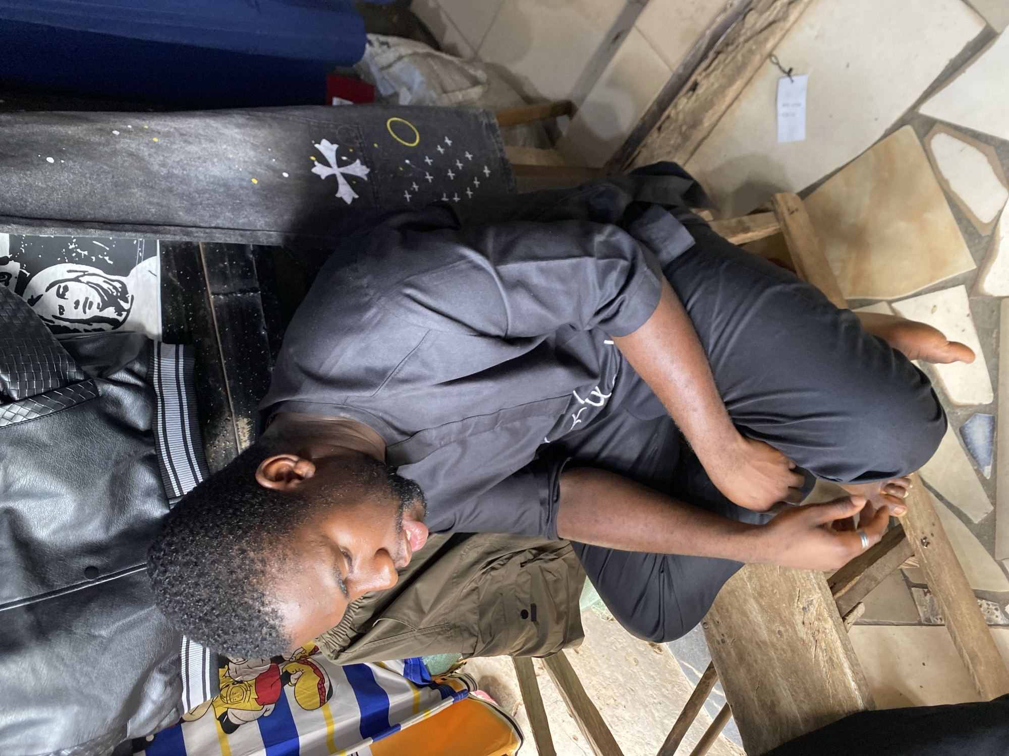A man is lying on a bench. He is wearing black clothing. The bench is located inside a building in Bukuru, Nigeria. The floor is tiled with mismatched tile pieces. The man's body is on a bench in a curled-up fetal position with his knees bent and pulled towards his chest. His arms are also bent. His eyes are closed. The bench is made of wooden planks. There is clothing under his body. There are other pieces of furniture in the scene.
