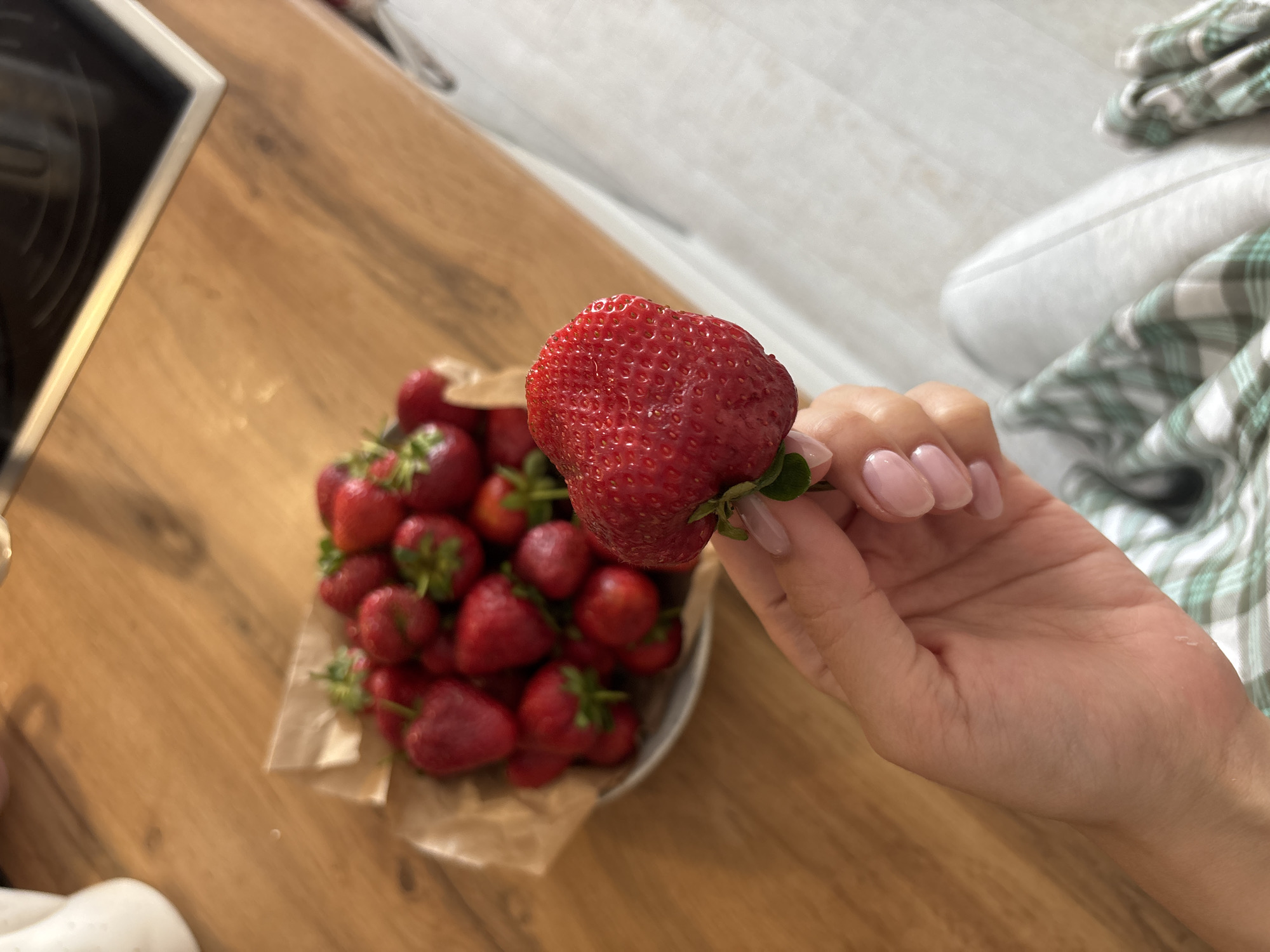 A photograph depicts a hand holding a large strawberry in front of a bowl filled with more strawberries. The bowl is placed on a wooden surface. The background includes part of a stove and a white and green plaid fabric. The location cannot be determined from the image.