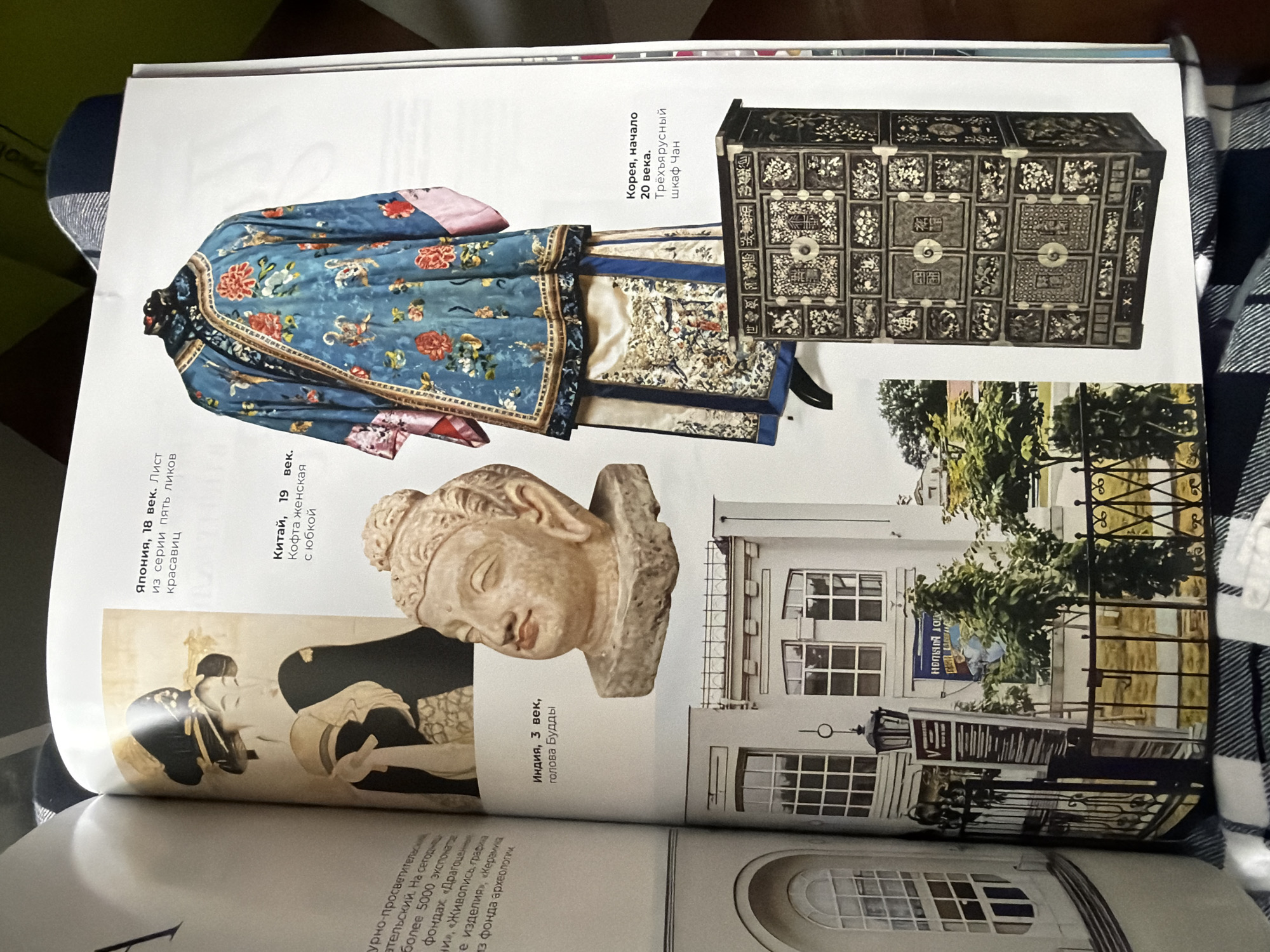 This is a photograph of a magazine page spread. The page displays images of cultural artifacts. On the left side, there's a depiction of a woman from 18th century Japan, a bust of Buddha from 3rd century India, and a woman's jacket from 19th century China. On the right, there is a blue embroidered jacket from Korea from the 20th century and a decorative chest, also from Korea. Below these artifacts is a photograph of the exterior of an art museum in Moscow, Russia. The museum building is white with multiple windows.