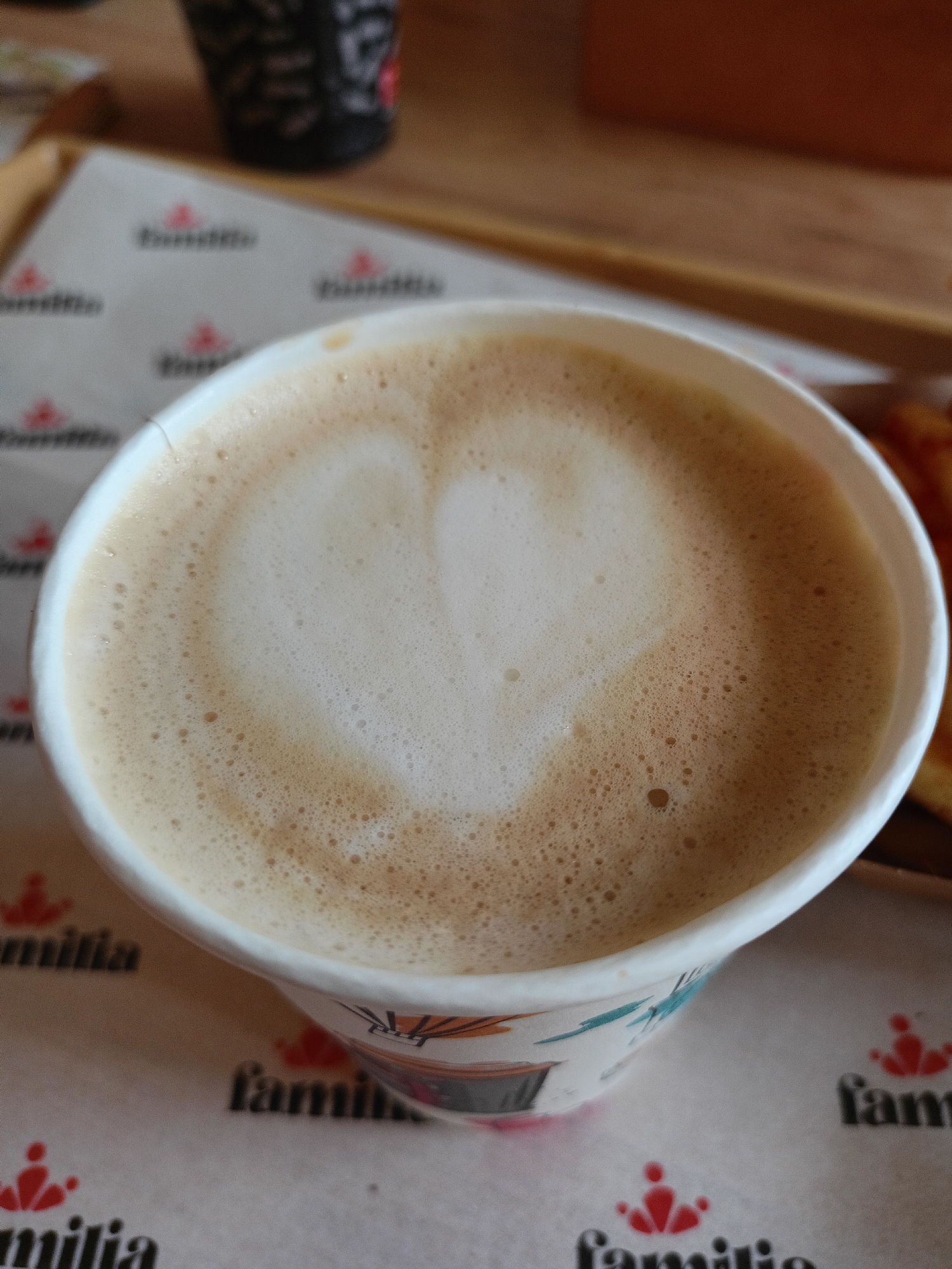 A close-up, overhead shot captures a white paper cup filled with a latte, featuring a heart shape artfully created in the foam. The cup is positioned on a surface covered with white paper bearing repeated "familia" logos and a small red icon. In the background, a wooden table and a blurred black cup are visible. The scene is likely set in a cafe or restaurant.