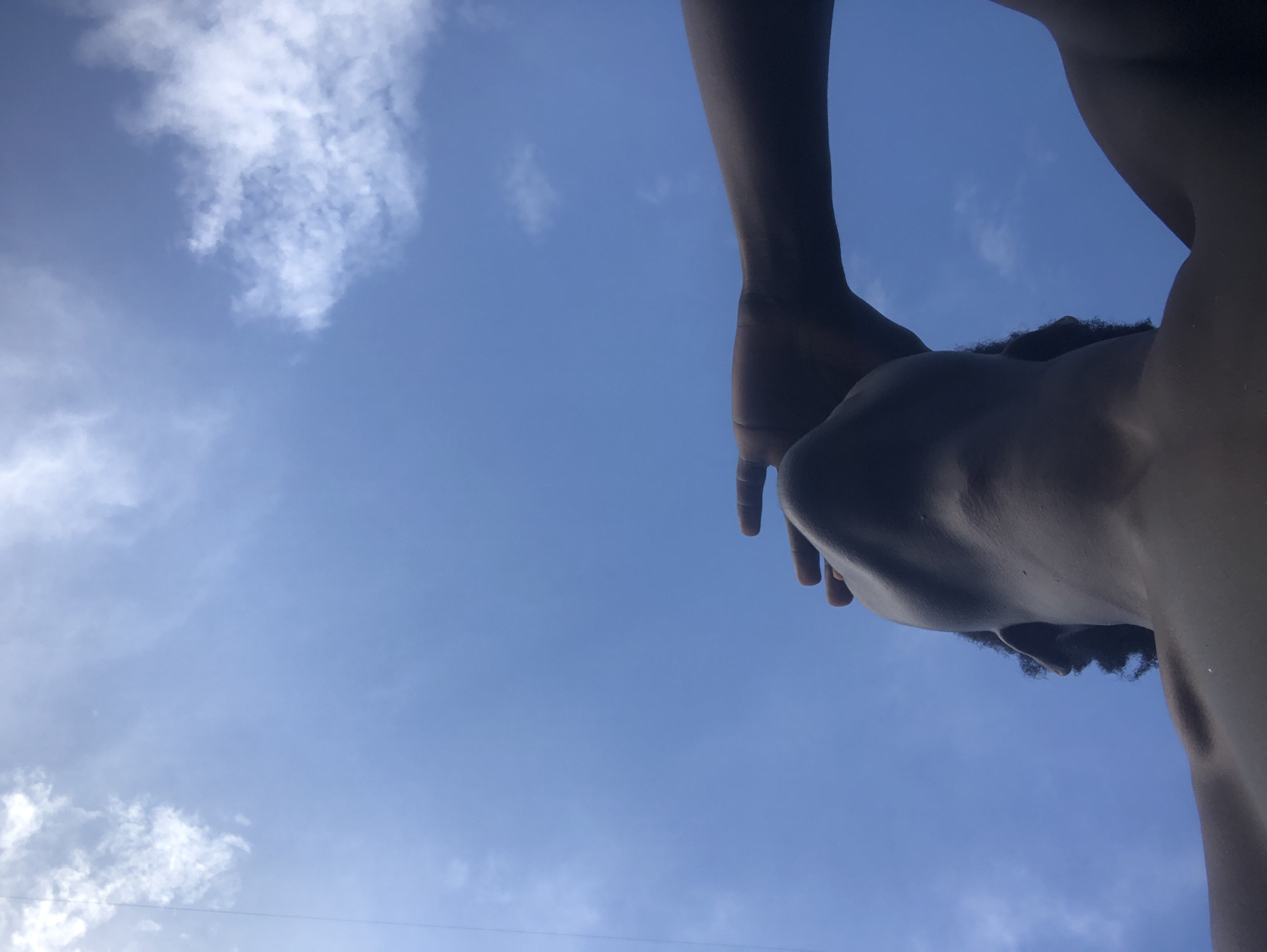 The image captures a low-angle shot of a person looking up at a bright blue sky with scattered white clouds. Their face is partially obscured by their hand, which is raised to shield their eyes from the sun. The person's neck and shoulders are visible, and they appear to be shirtless. The background is dominated by the expansive sky, suggesting an outdoor setting. The image does not provide any specific visual cues that would indicate a location within the city of Ado, Nigeria.