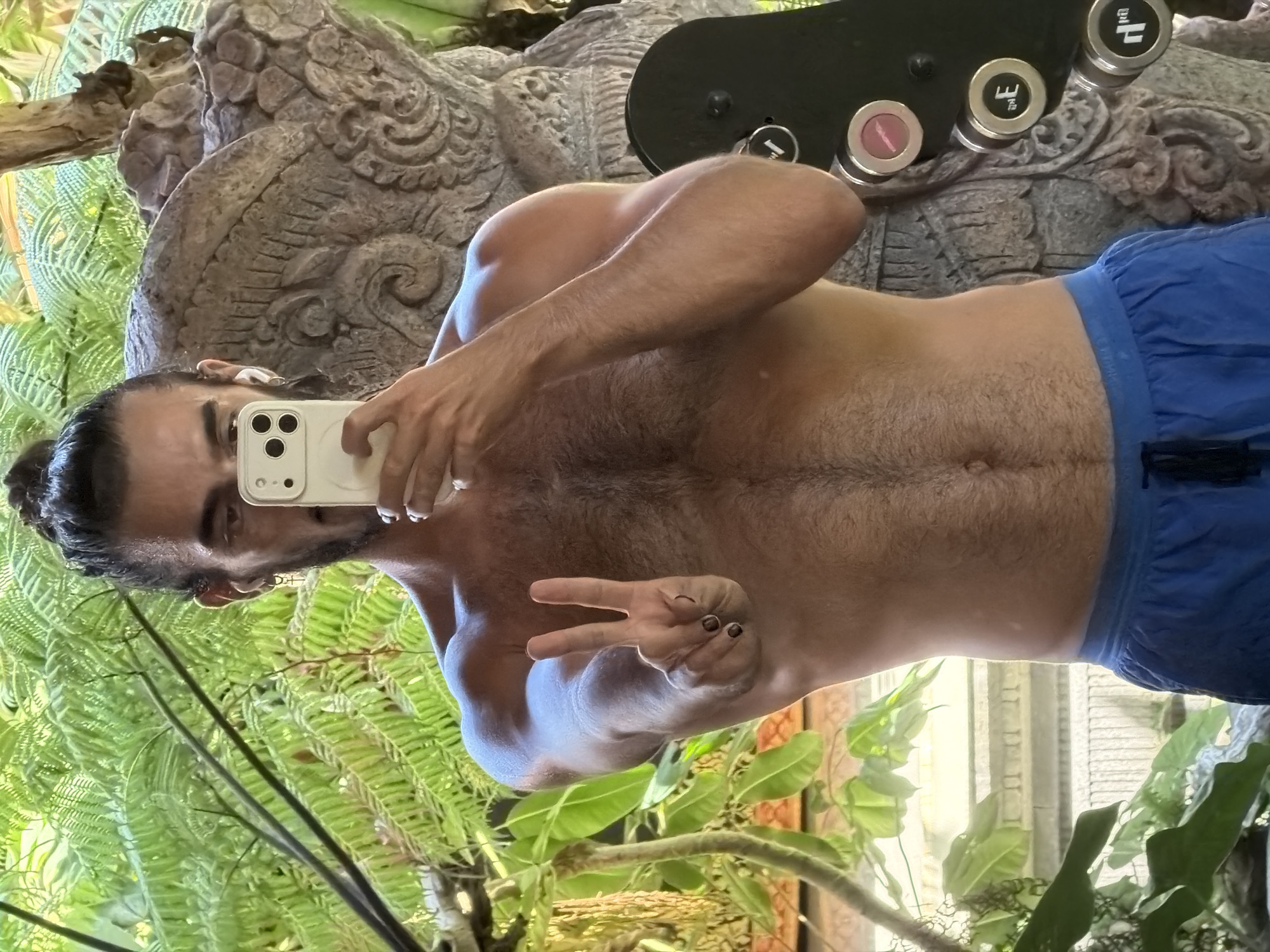 A shirtless man with dark hair pulled into a bun and a beard holds a smartphone up to take a selfie. He is wearing blue shorts and has a peace sign gesture with his right hand. The man is standing in front of lush green foliage and a stone carving. To the right of the man is a piece of gym equipment with weights. The scene appears to be an outdoor setting, possibly a garden or resort area in North Kuta, Indonesia.