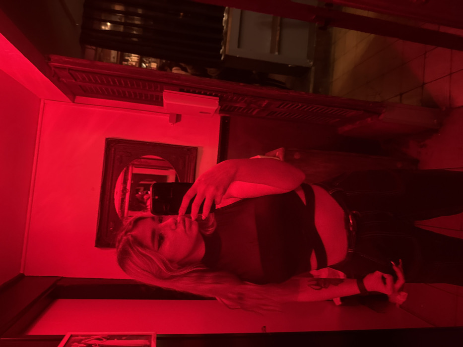 The image displays a mirror selfie of a young female subject within a red-lit interior, located in Tel-Aviv, Israel. The subject, possessing long, light-colored hair, prominent dark eye makeup, and a nose piercing, is dressed in a black, high-necked, sleeveless crop top with mesh panels and midriff cutouts, complemented by dark trousers and a belt. She is actively holding a black smartphone in her right hand to capture her reflection. The immediate environment includes a dark-framed mirror to the subject's right, reflecting a background suggestive of a bar or lounge, with shelves displaying bottles. To the subject's right, a partially open doorway, framed by what appears to be folded louvered doors, leads to a separate, dimly illuminated space also consistent with a bar setting. A white rectangular fixture, possibly a control panel, is mounted on the wall adjacent to the doorway. The entire scene is uniformly illuminated by intense red artificial lighting, imparting a strong reddish hue to all visible surfaces, including what appear to be light-colored walls and tiled flooring. A framed object is partially visible on the wall to the subject's left.