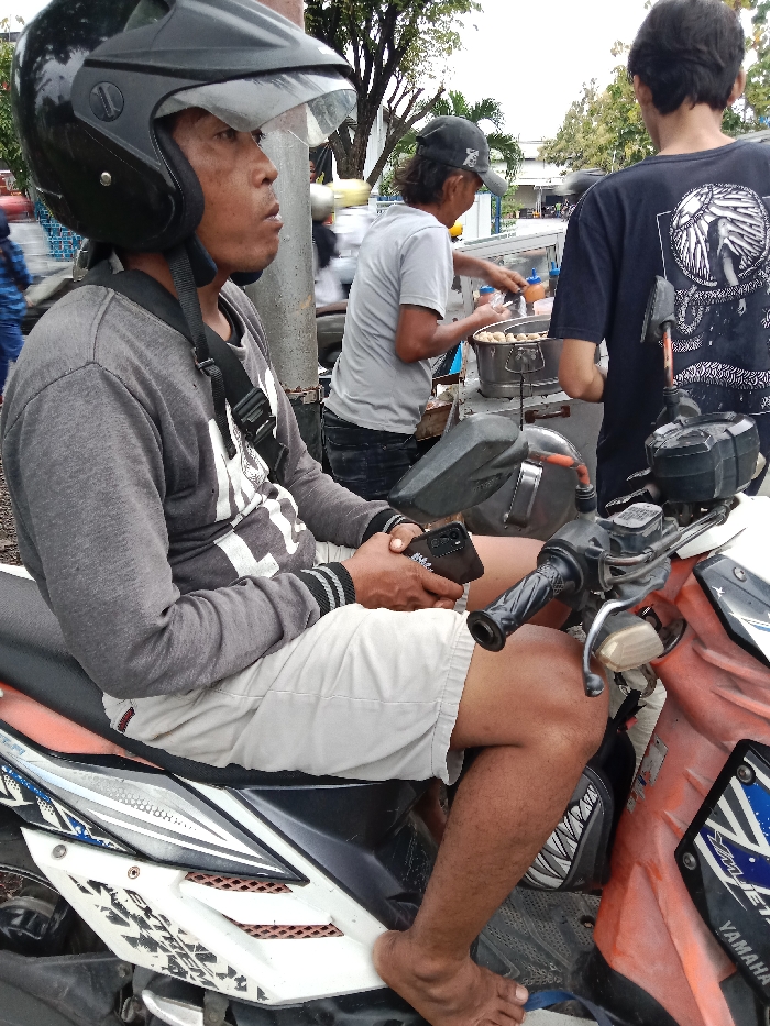 The image depicts a man wearing a motorcycle helmet and a grey sweater with a white graphic design, sitting on a white and orange motorcycle. He is holding a black smartphone in his right hand. A second man, wearing a grey shirt, is standing next to a food stall and appears to be interacting with another man in a black shirt. The image is taken on a street in Tugu, Indonesia, located at coordinates -6.973 latitude and 110.326 longitude.