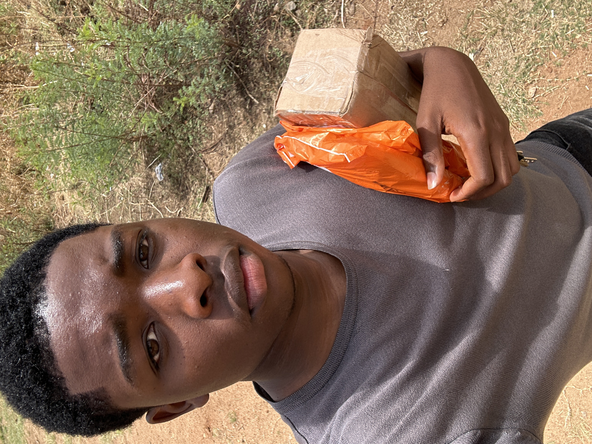 A young man is holding a small cardboard box and an orange plastic bag. He is standing outdoors in a location with dry grass and a small green bush. It is daytime, and the light is bright. The city, Kirana, Nigeria, cannot be confirmed by the image.