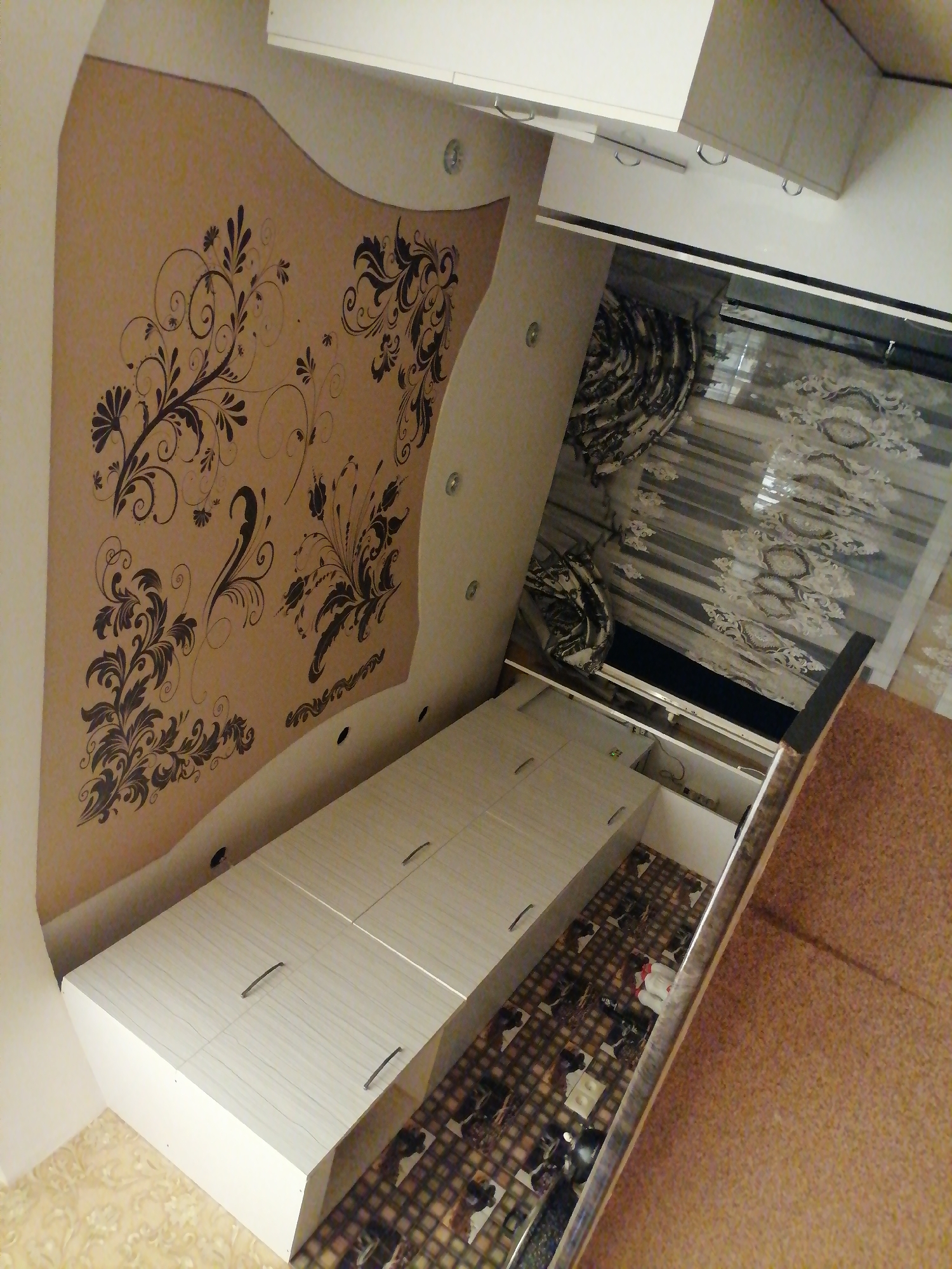 The image depicts an interior scene that appears to be a bedroom or a living area with built-in furniture. The dominant elements are a large, white, modular cabinet unit that takes up a significant portion of the foreground and extends into the mid-ground. This cabinet has multiple drawers and what appears to be a pull-out section. Above the cabinet, a patterned wallpaper or mural with dark floral motifs is visible on a curved wall. To the right of the cabinet, a window is covered by blinds with an intricate, possibly embroidered, fabric overlay. The floor has a checkered pattern with images and appears to be carpeted in some areas, particularly in the far right of the image. The overall lighting suggests it is daytime, but the room itself is not brightly lit. There are no people visible in the image. The style of the furniture and decor suggests a modern, possibly custom-designed space. The mention of Rustavi, Georgia, in the prompt suggests this is a residential interior located in that city.