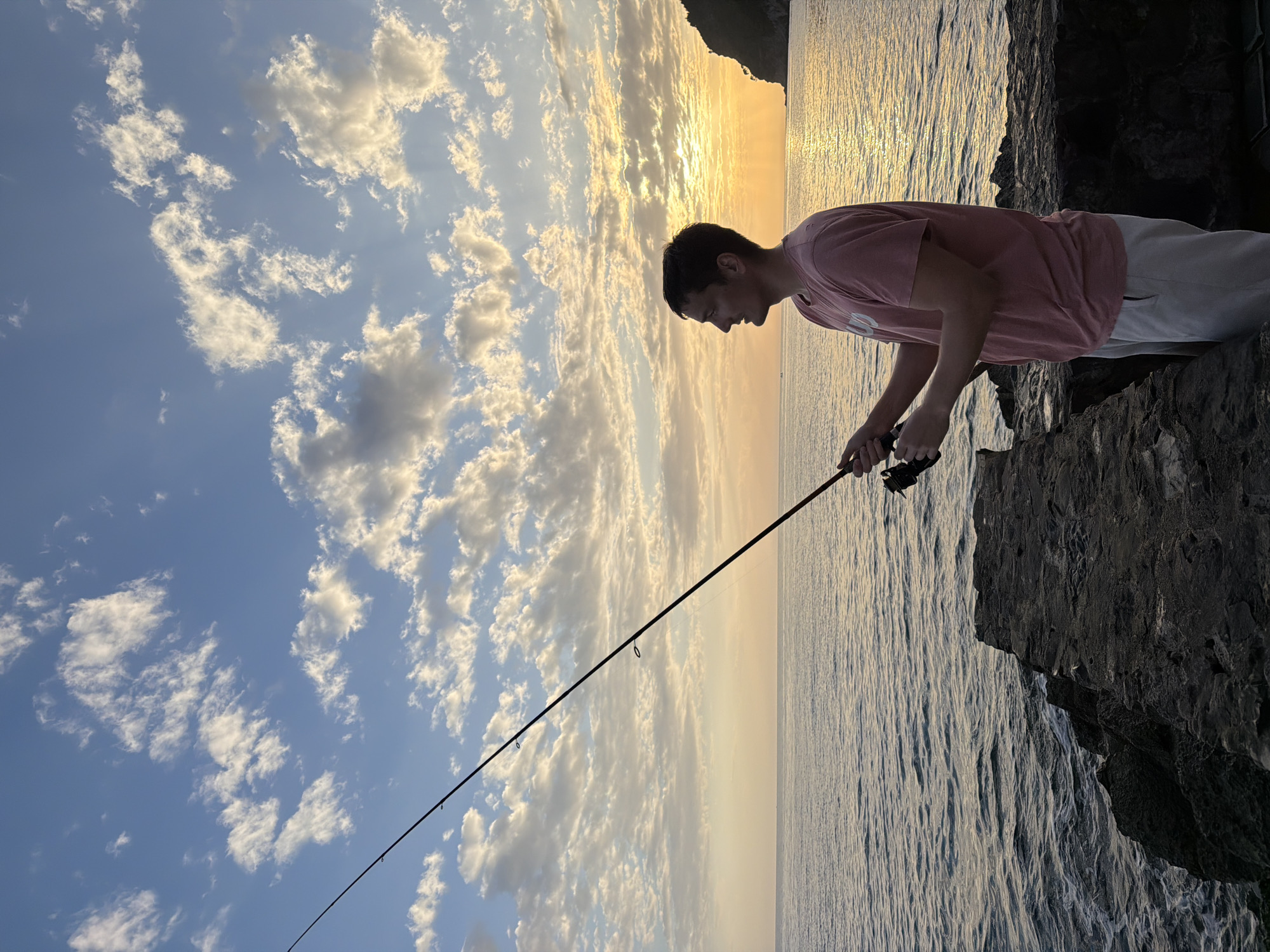 A man is fishing off a rocky cliff in Petrovac centar, Montenegro. He is wearing a pink t-shirt and light-colored pants. He holds a fishing rod and looks down at the water. The sky is blue with scattered white clouds, and the sun is setting over the water, creating a golden glow. The sea is calm with gentle ripples. A dark, rocky coastline is visible in the background.