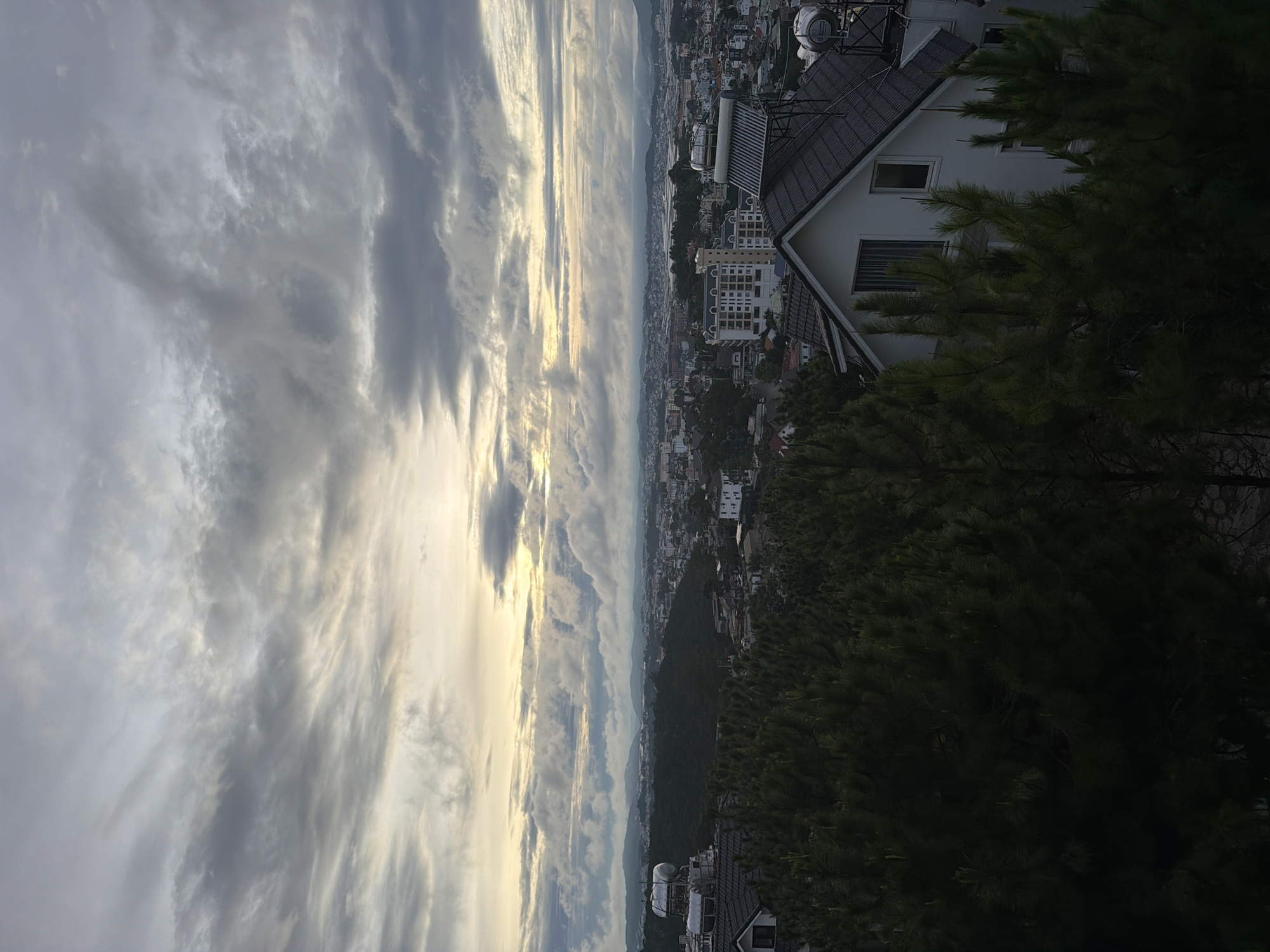 This image is an outdoor, daytime, full shot of a city skyline from an elevated viewpoint. The sky is overcast with dramatic clouds and a hint of golden sunlight breaking through. In the foreground, dense green pine trees partially obscure a white house with a dark tiled roof, featuring a solar water heater on the roof. Beyond the trees, the city of Xuân Trường - Đà Lạt, Vietnam, stretches out, characterized by a mix of modern buildings and residential houses, nestled among rolling hills and lush greenery. The city appears quiet and undeveloped, with a vast expanse of trees dominating the landscape.