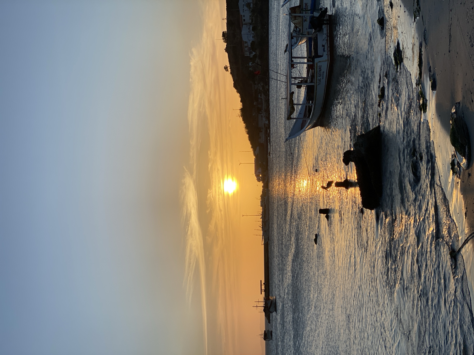 The image depicts a sunset over the water in Juan Griego, Venezuela. The sun is low on the horizon, casting a golden glow across the sky and reflecting on the water's surface. A small boat is anchored near the shore. The shoreline consists of sand and some rocks, with some visible algae. A hillside with buildings is partially visible in the background. The sky transitions from blue to orange as the sun approaches the horizon.